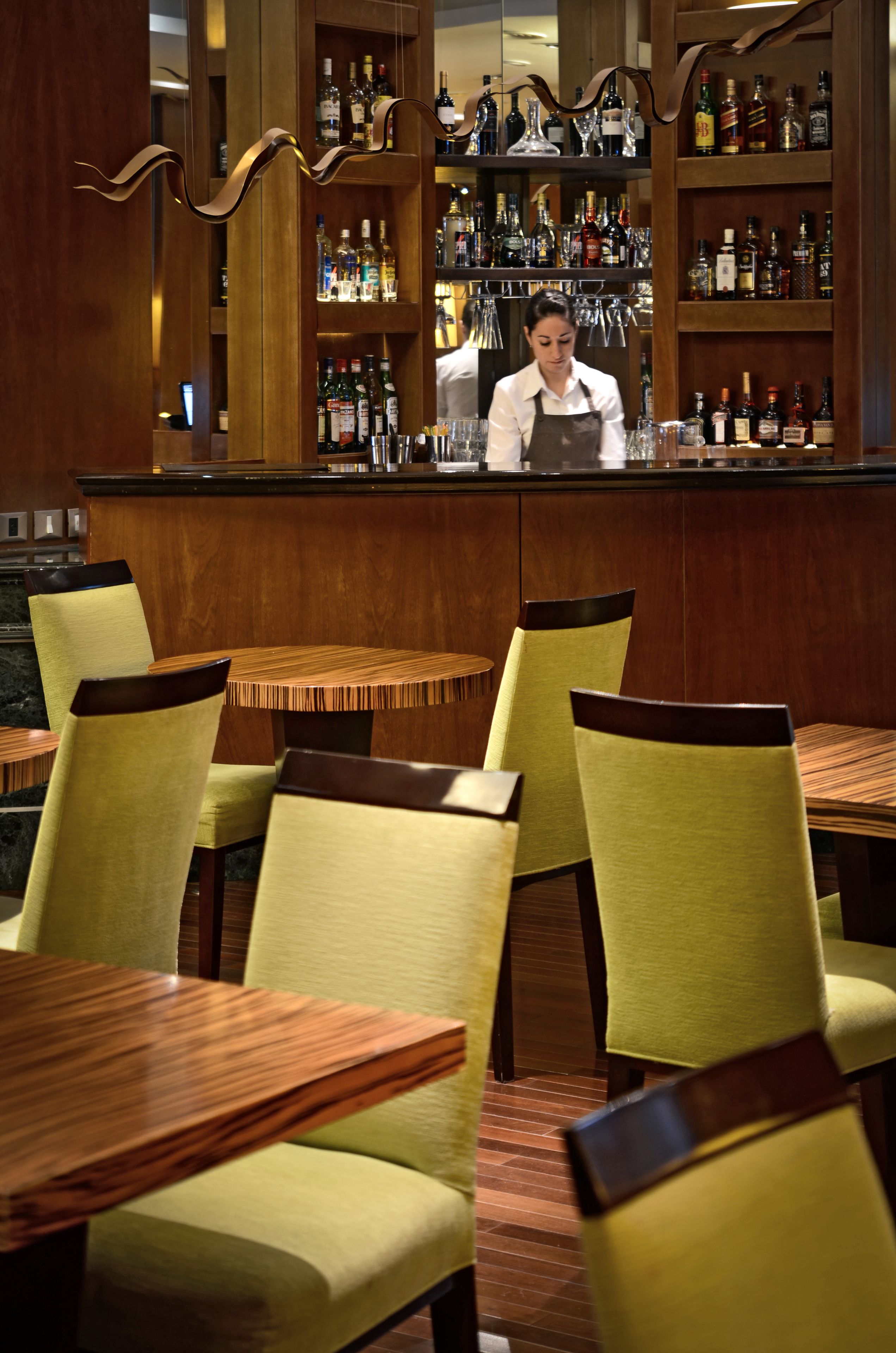 A bar area with wooden shelves stocked with bottles and glasses, a bartender at the counter, and green upholstered chairs around wooden tables.
