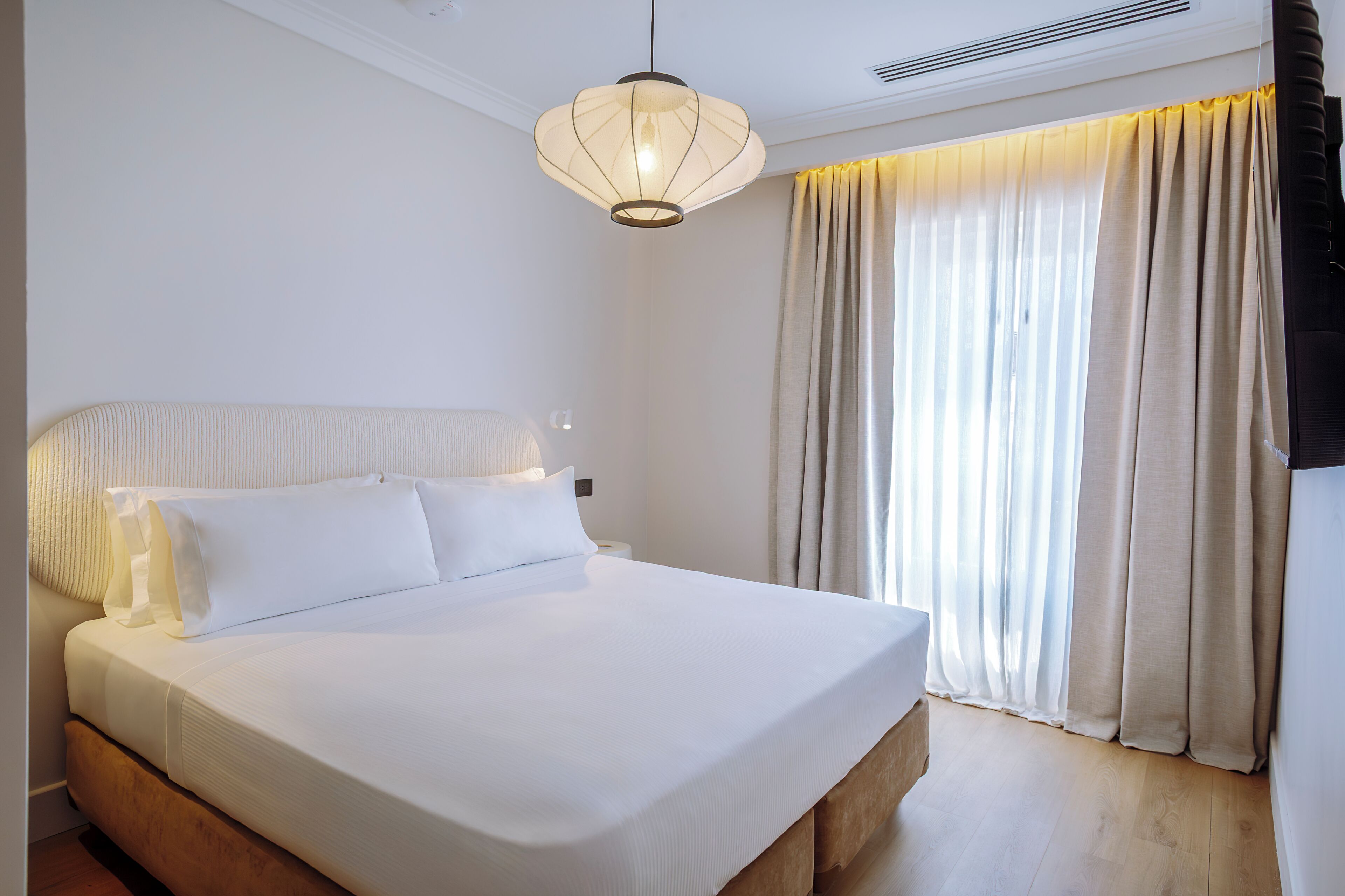 Een net opgemaakt bed met witte lakens, een hanglamp en een raam met gordijnen.