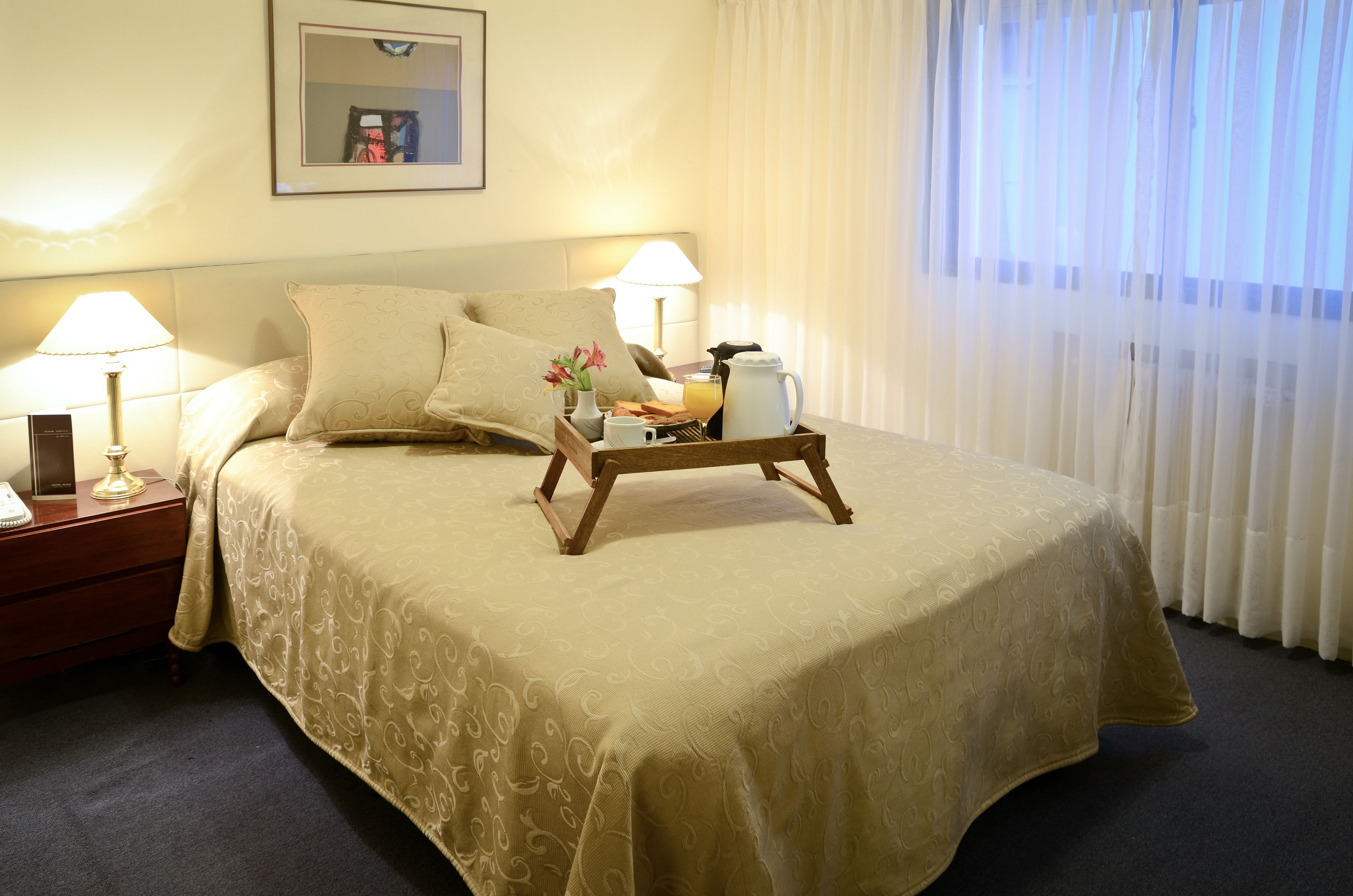 Een keurig opgemaakt bed met een beige sprei, twee nachtlampjes, een dienblad met ontbijtartikelen en een lijst aan de muur.