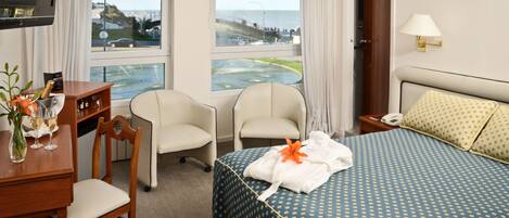 A hotel room with a bed, two chairs, a desk with a vase of flowers, a lamp, and a view of a body of water and a bridge.