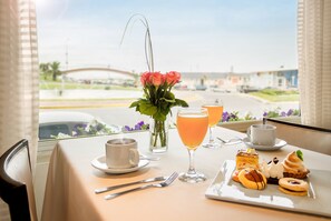 Ein Esstisch mit einer Tasse, einem Glas Orangensaft, einem Glas Weißwein, einem Teller mit Gebäck und einer Vase mit Blumen.