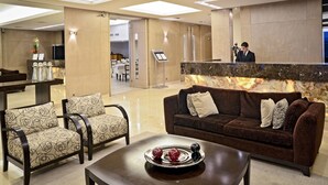A lobby with a reception desk, a brown sofa, two patterned armchairs, a coffee table with decorative items, and a large mirror. - Hotel Iruña (Mar del Plata)