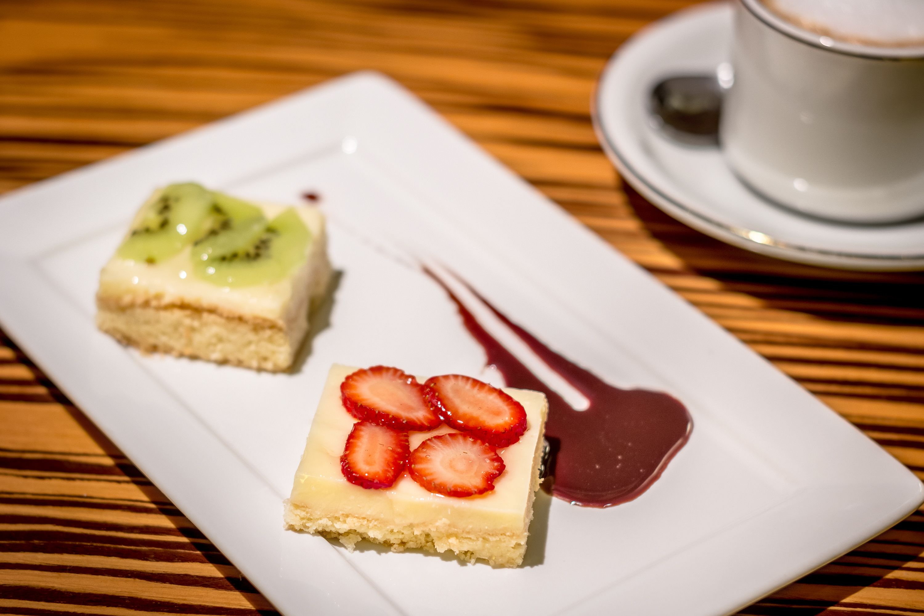 A dessert plate with two square pieces of cake, one topped with kiwi and the other with strawberries, and a cup of coffee on a saucer.