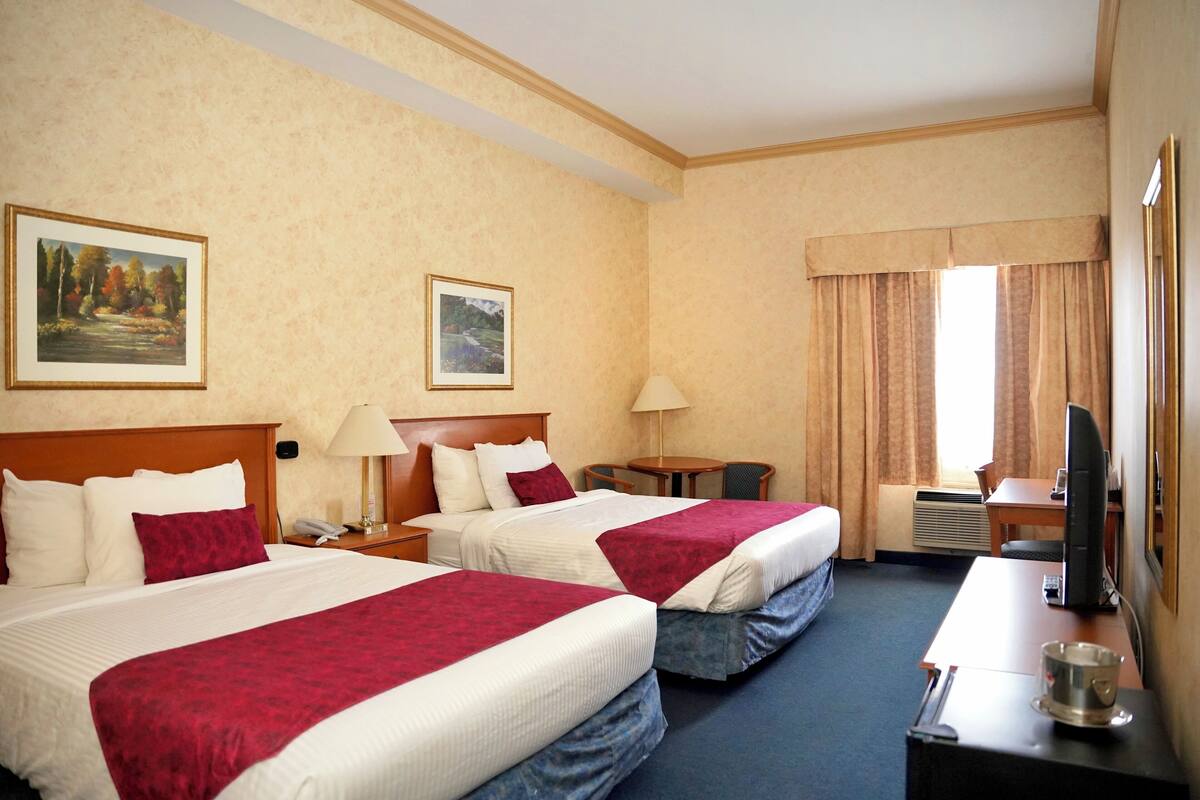 A hotel room with two beds, a desk with a TV, a chair, a table, and a window with curtains.