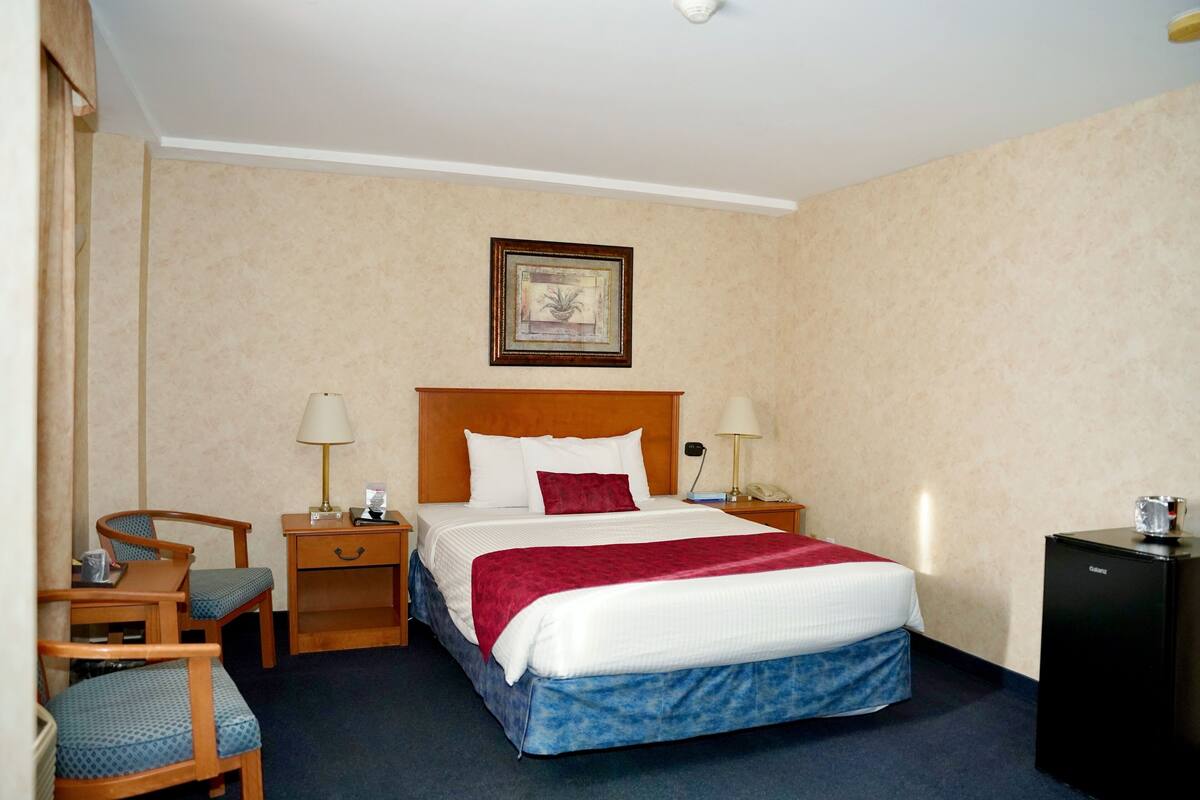 A hotel room with a bed, two bedside tables, a desk with a chair, a small refrigerator, and a framed picture on the wall.