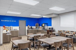 A breakfast area with tables, chairs, a kitchenette, and a blue wall with a 'Breakfast is on us' sign. - Days Inn by Wyndham Leamington (Leamington)