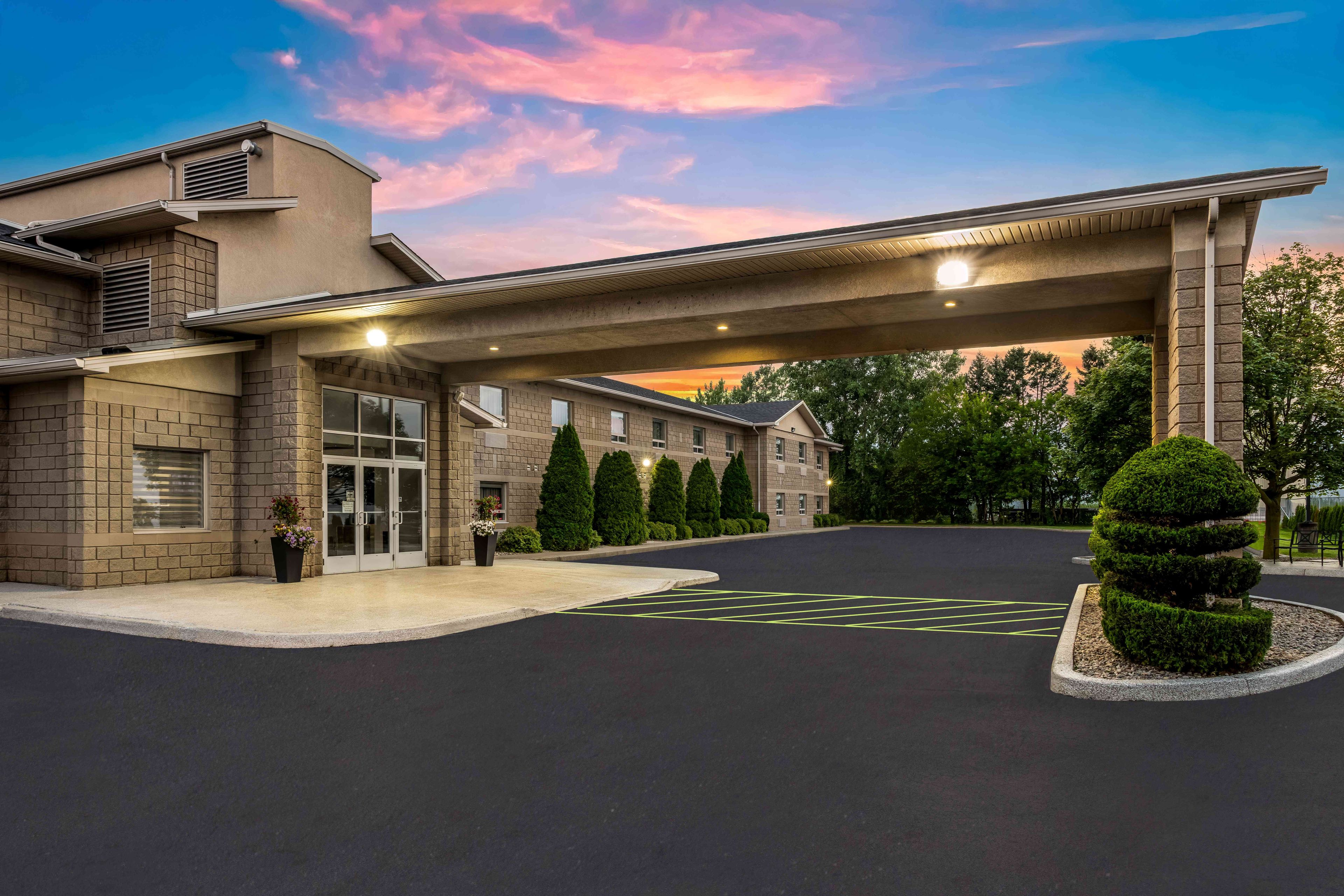 A hotel entrance with a covered driveway, a parking lot, and a landscaped area with trimmed bushes.