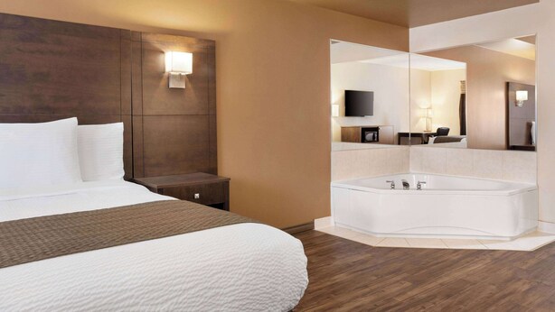 A hotel room with a large bed, a wooden headboard, a nightstand, a wall-mounted TV, a bathroom with a bathtub, and a mirror. - Days Inn by Wyndham Leamington (Leamington)