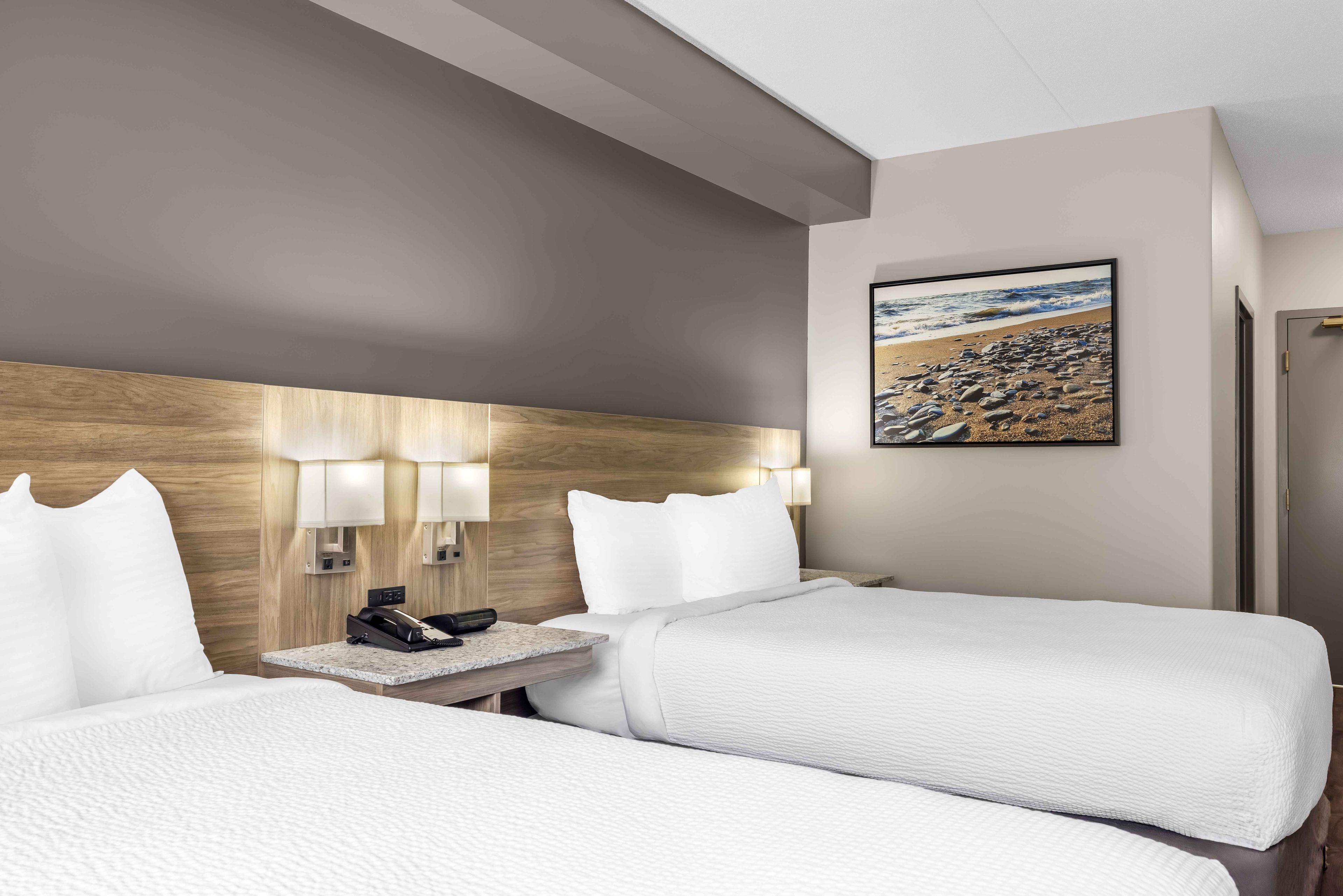 Habitación de hotel con dos camas, cabecera, mesita de noche con teléfono y lámpara, y una pintura de una playa.