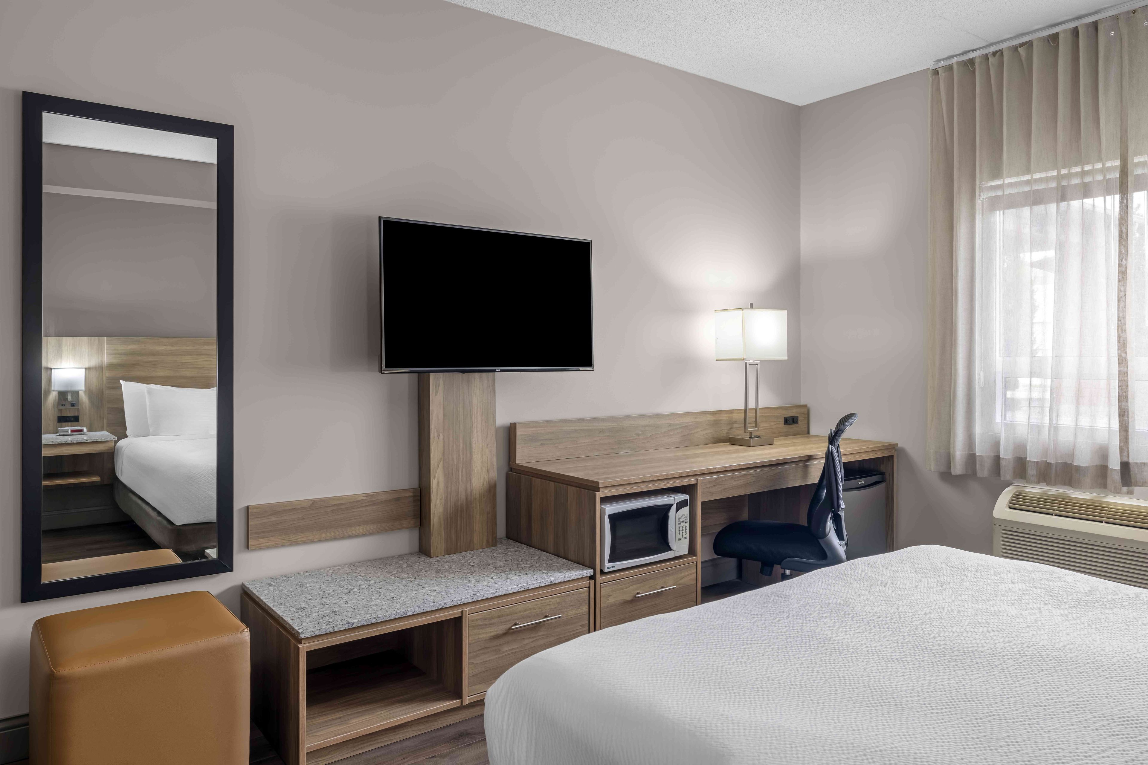 Ein Hotelzimmer mit einem Bett, einem Schreibtisch mit Stuhl, einem Fernseher, einem Spiegel, einer Lampe und einem Fenster mit Vorhängen.