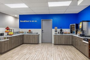 Un espace réservé au petit-déjeuner avec un mur bleu, une pancarte indiquant « Le petit-déjeuner est offert » et divers distributeurs de nourriture et de boissons.