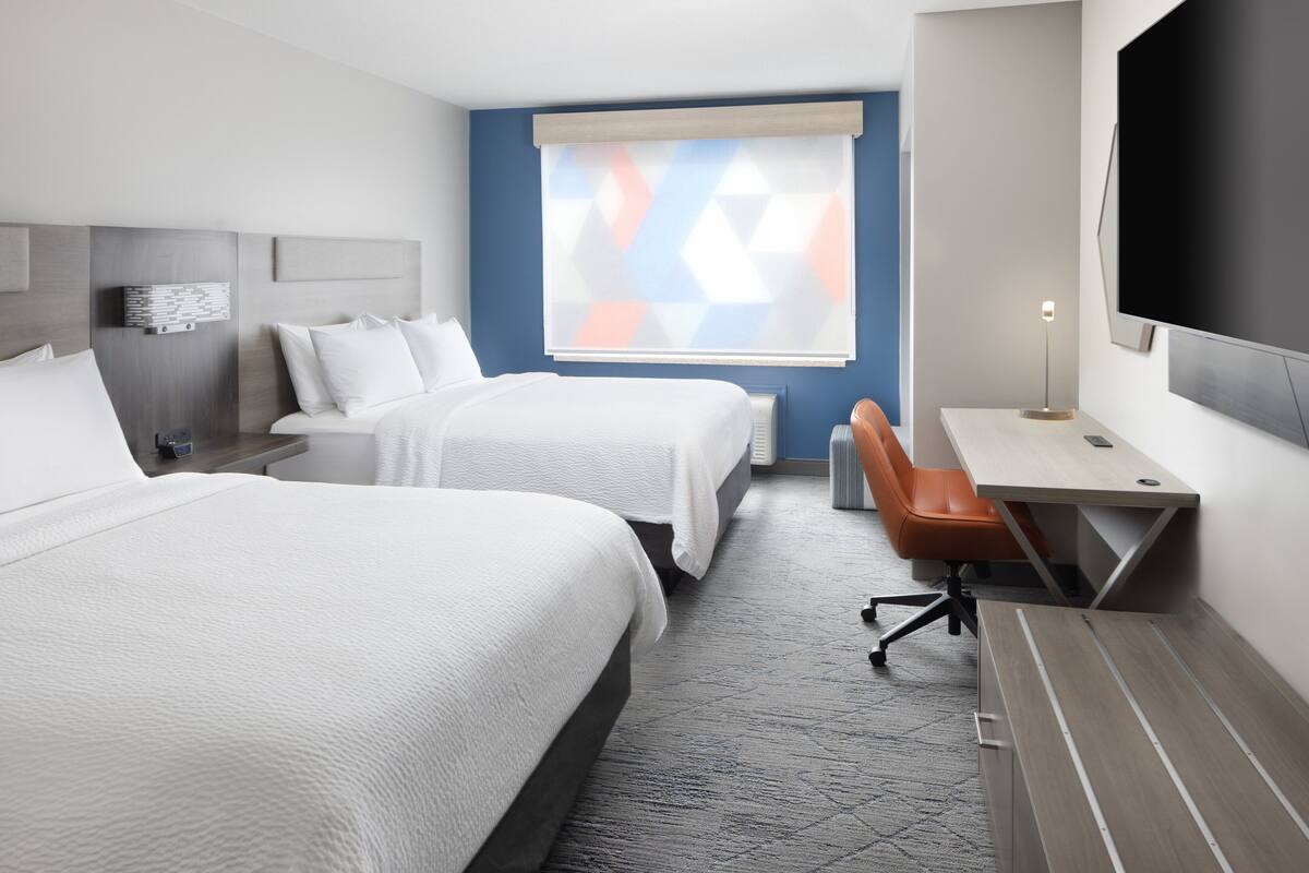 A hotel room with two beds, a desk, a chair, a TV, and a window with blinds.