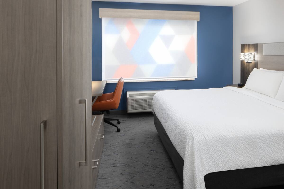 A hotel room with a bed, a desk, an orange chair, a window with a geometric pattern, and a nightstand with a lamp.