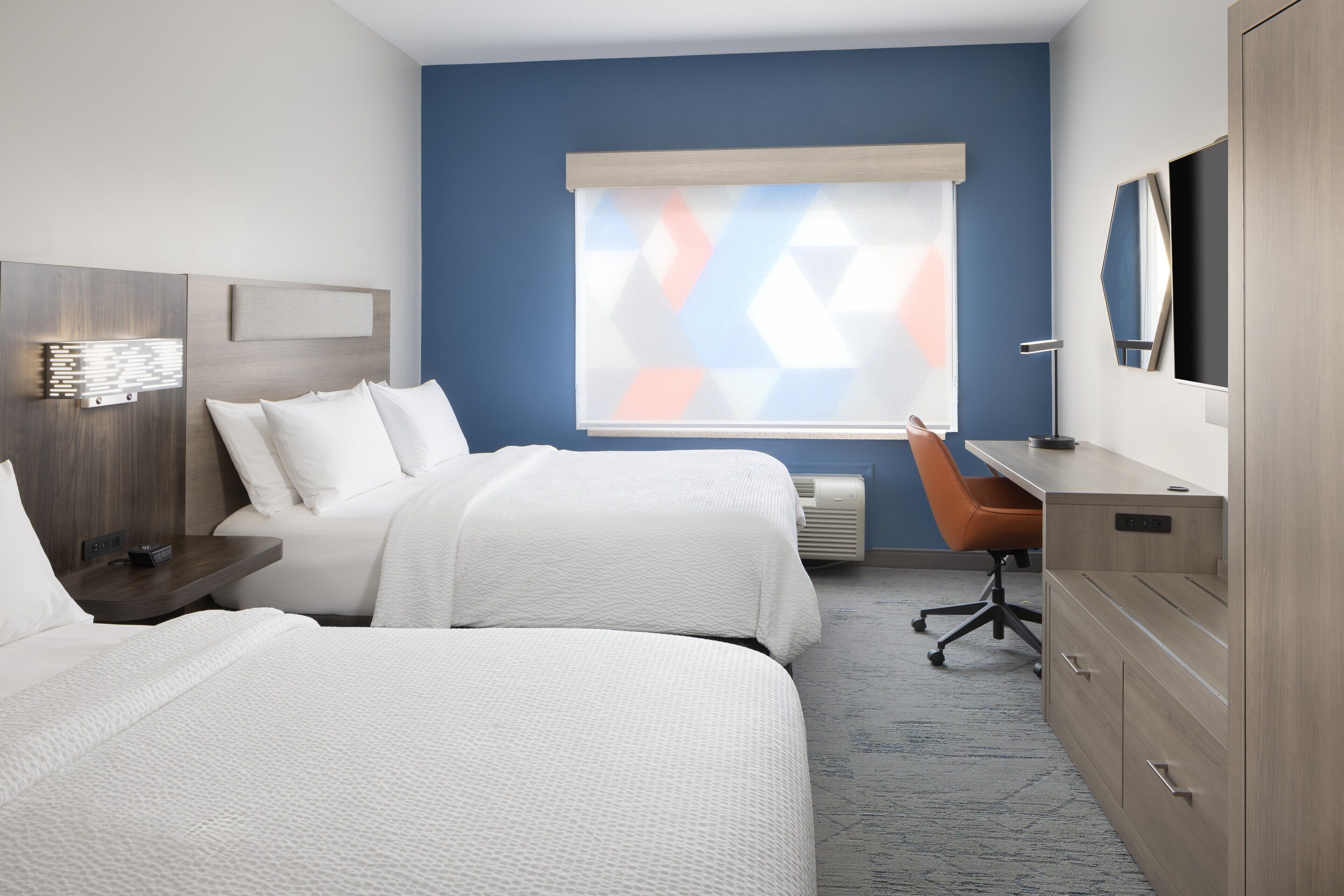 A hotel room with two beds, a desk, a chair, a TV, and a window with blinds.