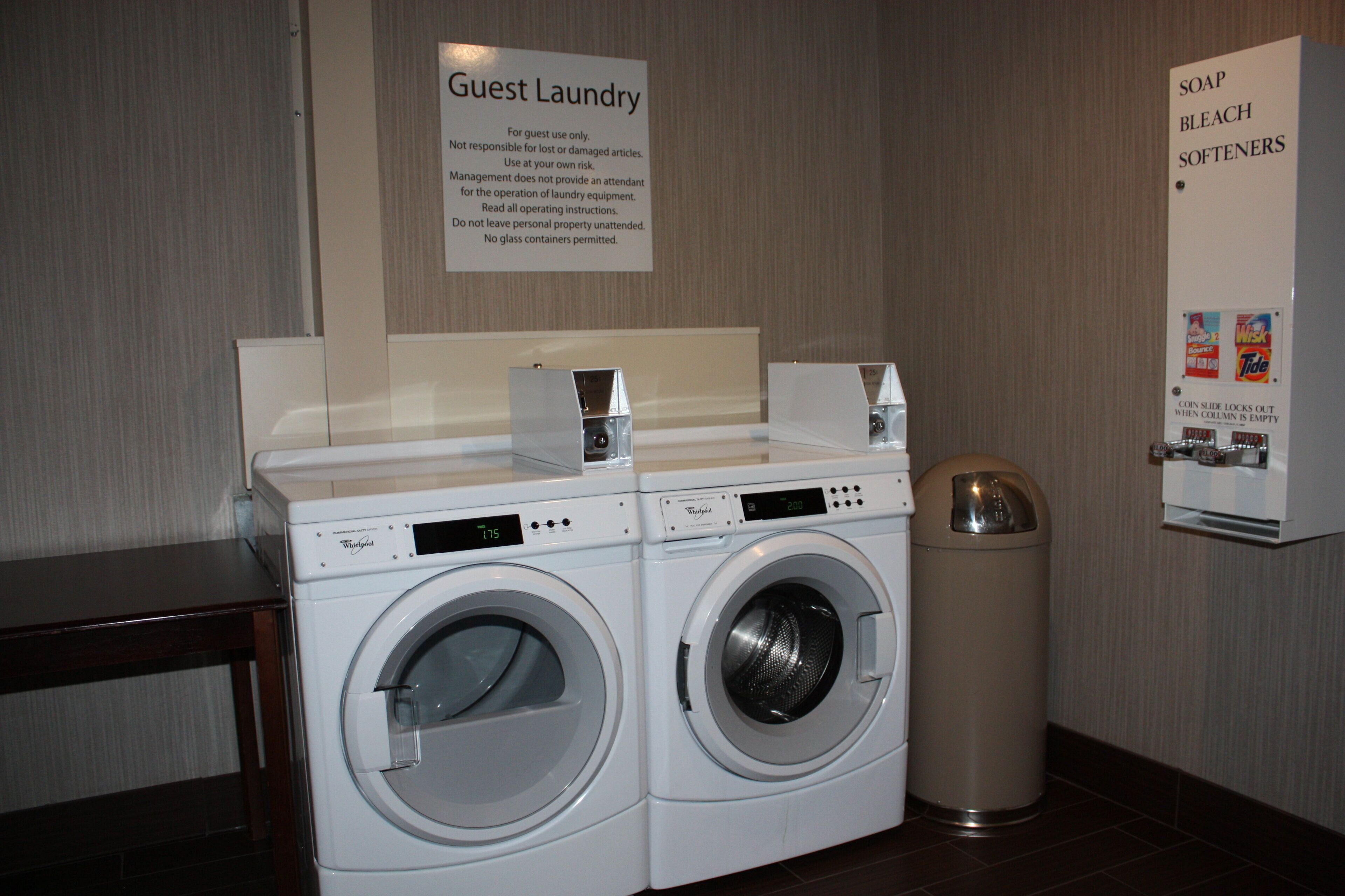 A guest laundry area with two Whirlpool washers, a coin-operated dryer, and a sign indicating the use of soap, bleach, and softeners.