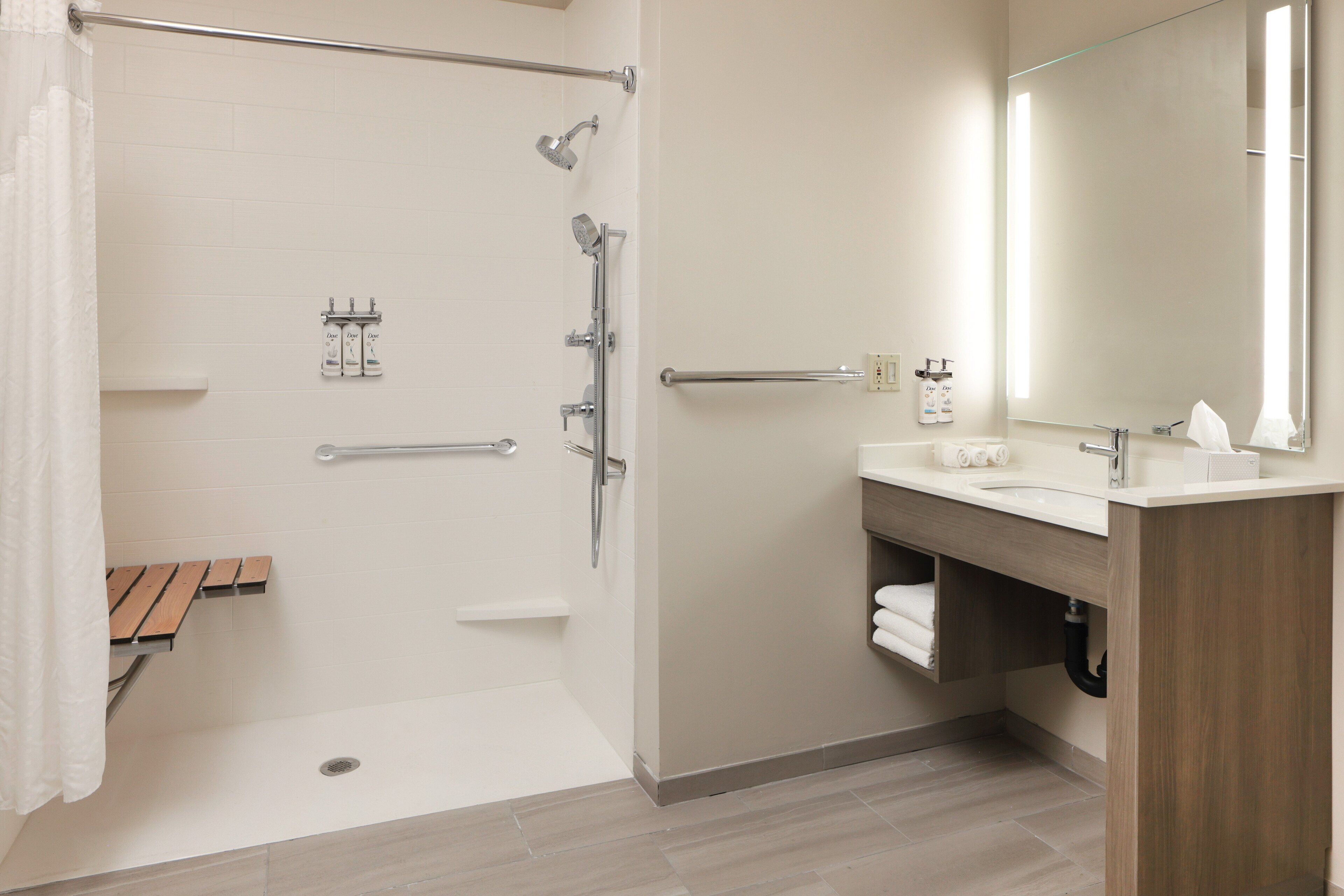A modern bathroom with a shower, grab bars, a wooden bench, a sink, and a mirror.