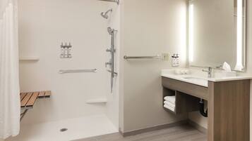 A modern bathroom with a shower, grab bars, a wooden bench, a sink, and a mirror.