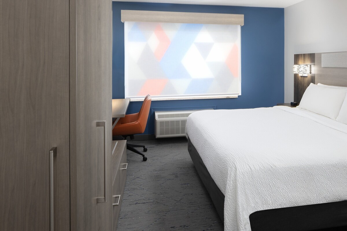 A hotel room with a bed, a desk, an orange chair, a window with a geometric pattern, and a nightstand with a lamp.