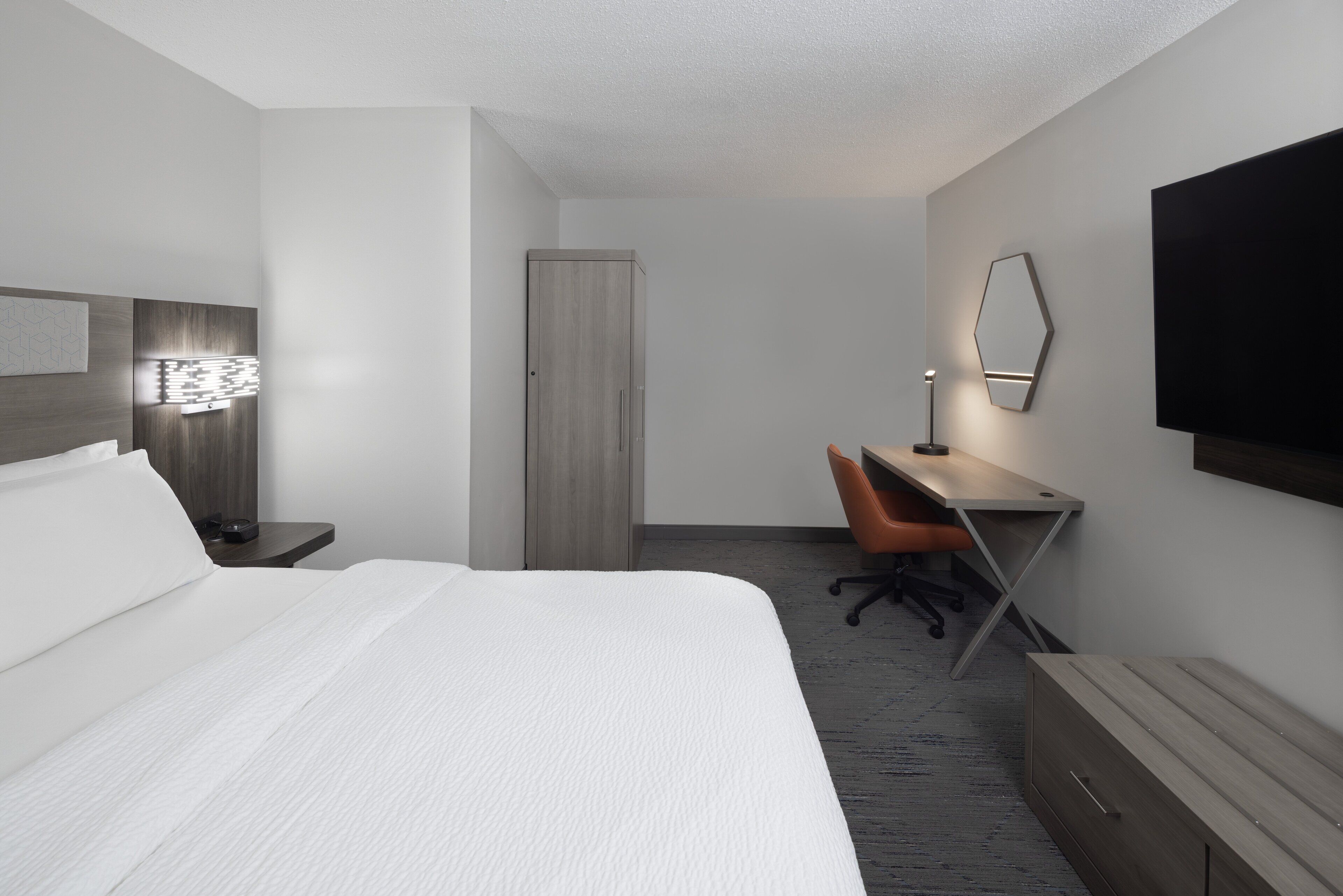 Una habitación de hotel moderna con una cama grande, un escritorio, una silla de oficina, un armario, un televisor montado en la pared y un espejo.
