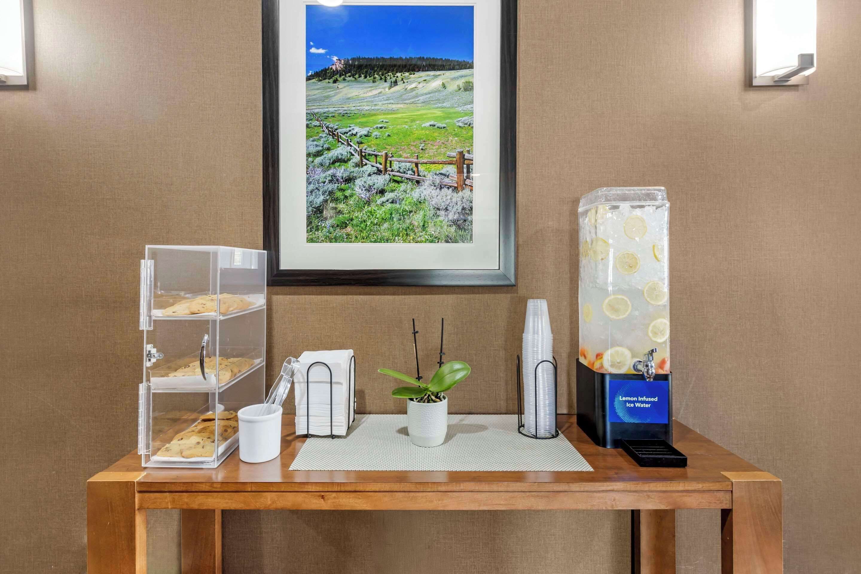 A wooden table with a framed landscape photo, a glass display case with pastries, a white mug, a potted plant, and a lemon-infused water dispenser.