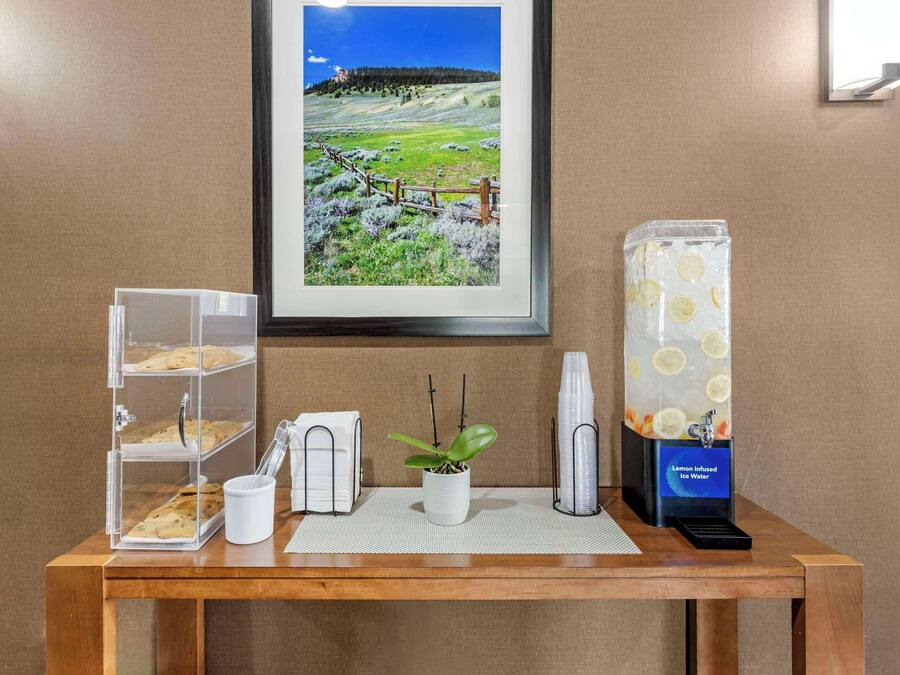 A wooden table with a framed landscape photo, a glass display case with pastries, a white mug, a potted plant, and a lemon-infused water dispenser.