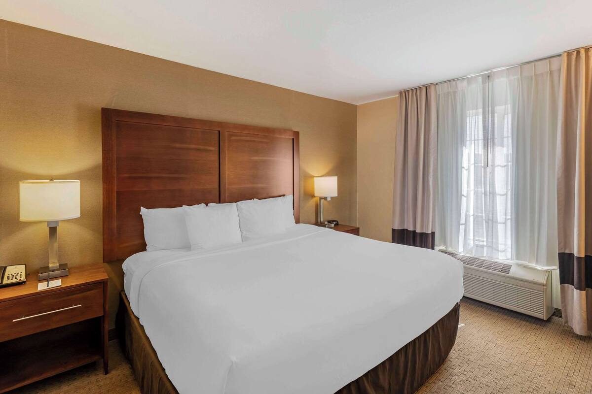 Una habitación de hotel con una cama grande, dos mesitas de noche con lámparas, una ventana con cortinas y un aire acondicionado instalado en la pared.