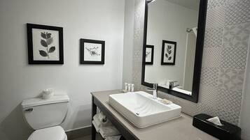 A modern bathroom with a toilet, sink, and large mirror. There are framed pictures on the wall and a towel rack.