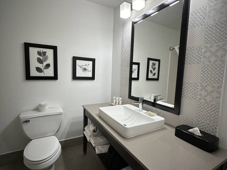 A modern bathroom with a toilet, sink, and large mirror. There are framed pictures on the wall and a towel rack.