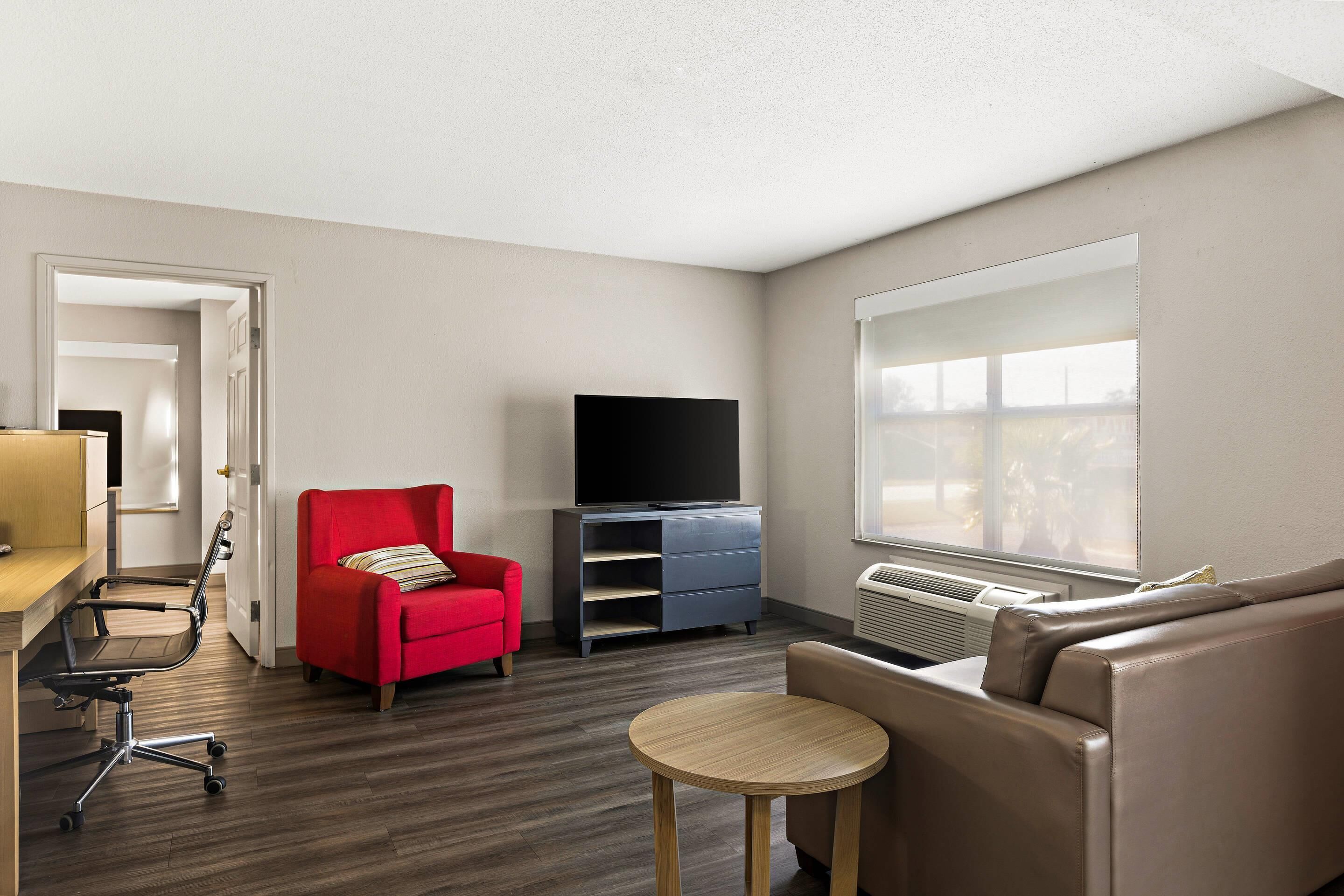 A modern living room with a flat-screen TV, a red armchair, a beige sofa, a small round table, and a wooden floor.