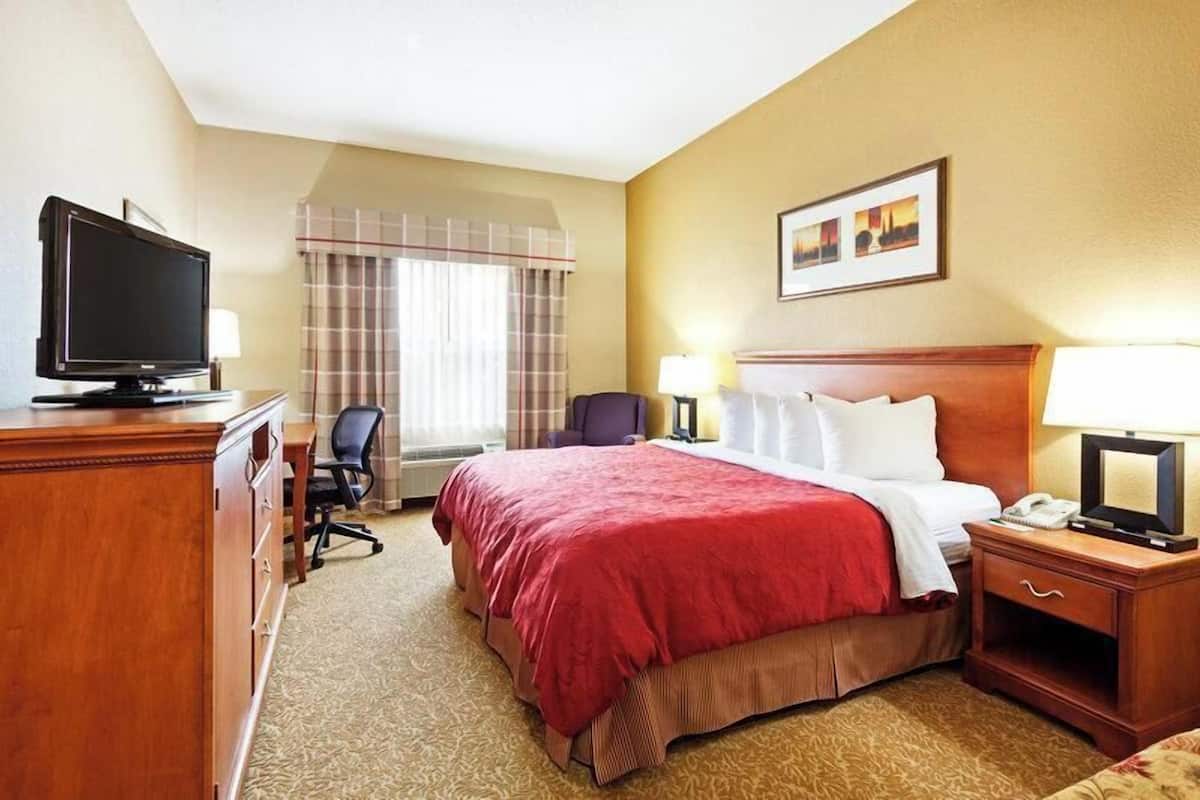 A hotel room with a bed, a desk with a television, a chair, a nightstand with a lamp, and a window with curtains.