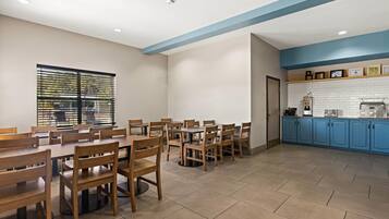 Uma sala de jantar com mesas e cadeiras de madeira, uma cozinha com armários azuis e uma janela com persianas.