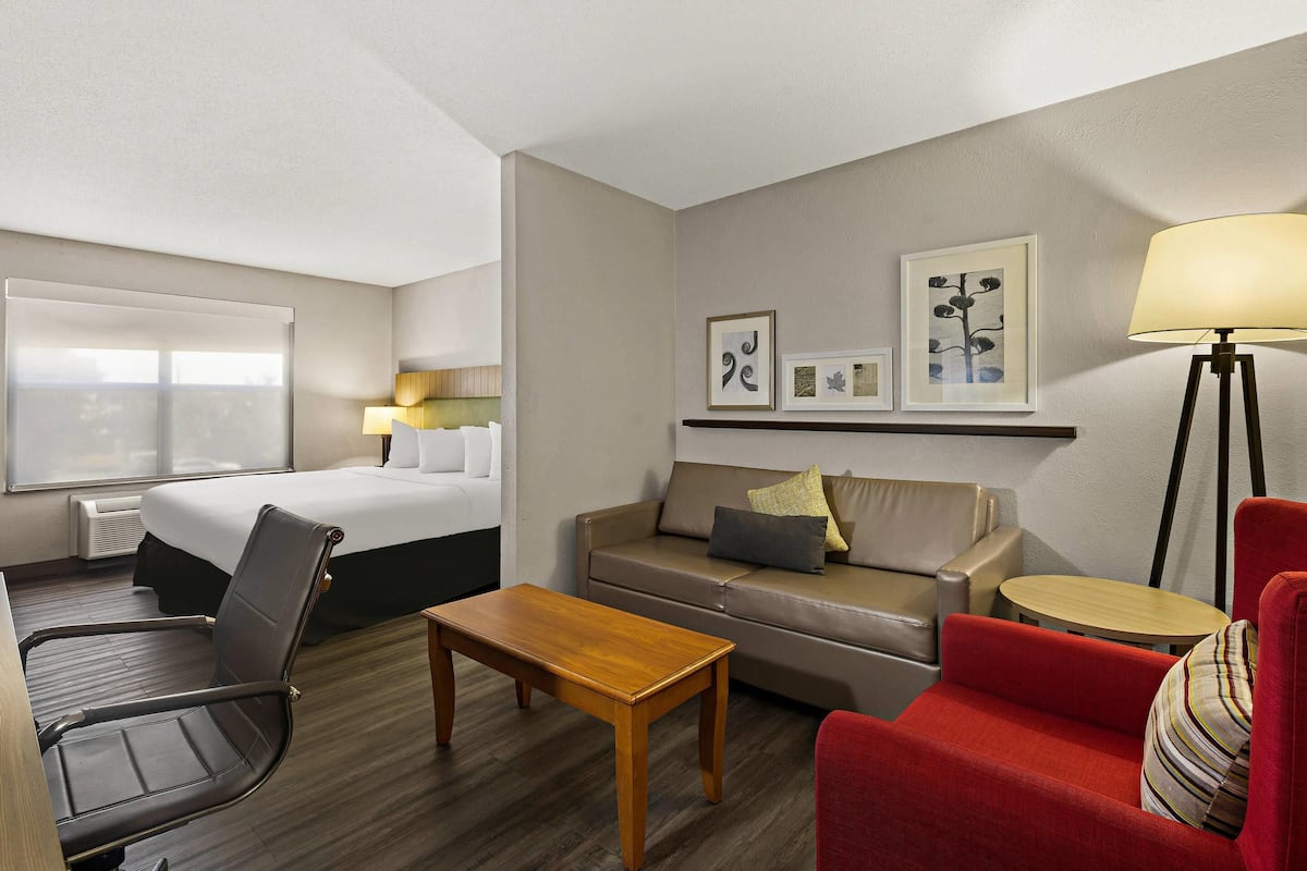 A hotel room with a bed, a sofa, a red armchair, a wooden coffee table, a floor lamp, and wall art.