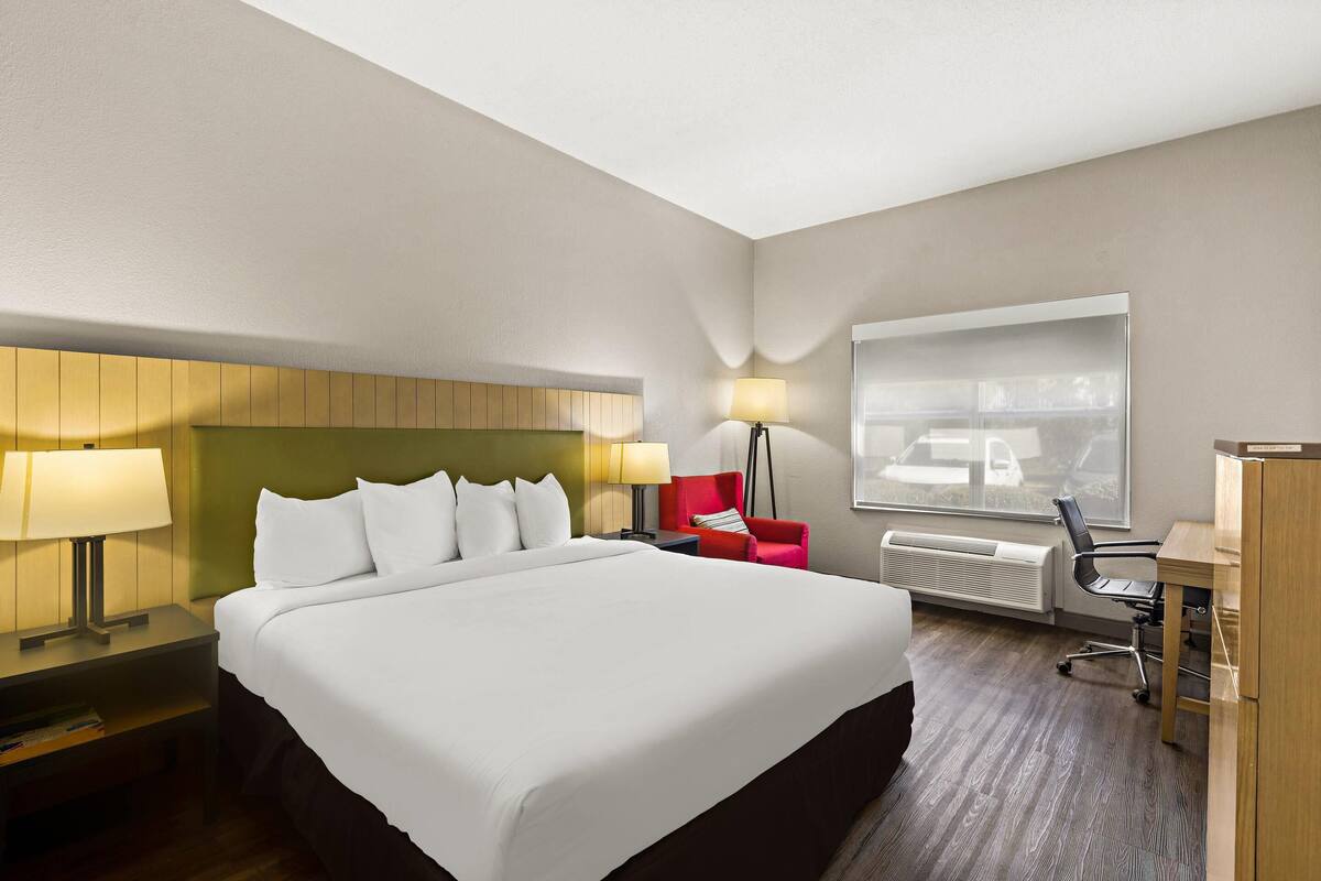A hotel room with a large bed, a desk with a chair, a red chair, two lamps, and a window with a view.