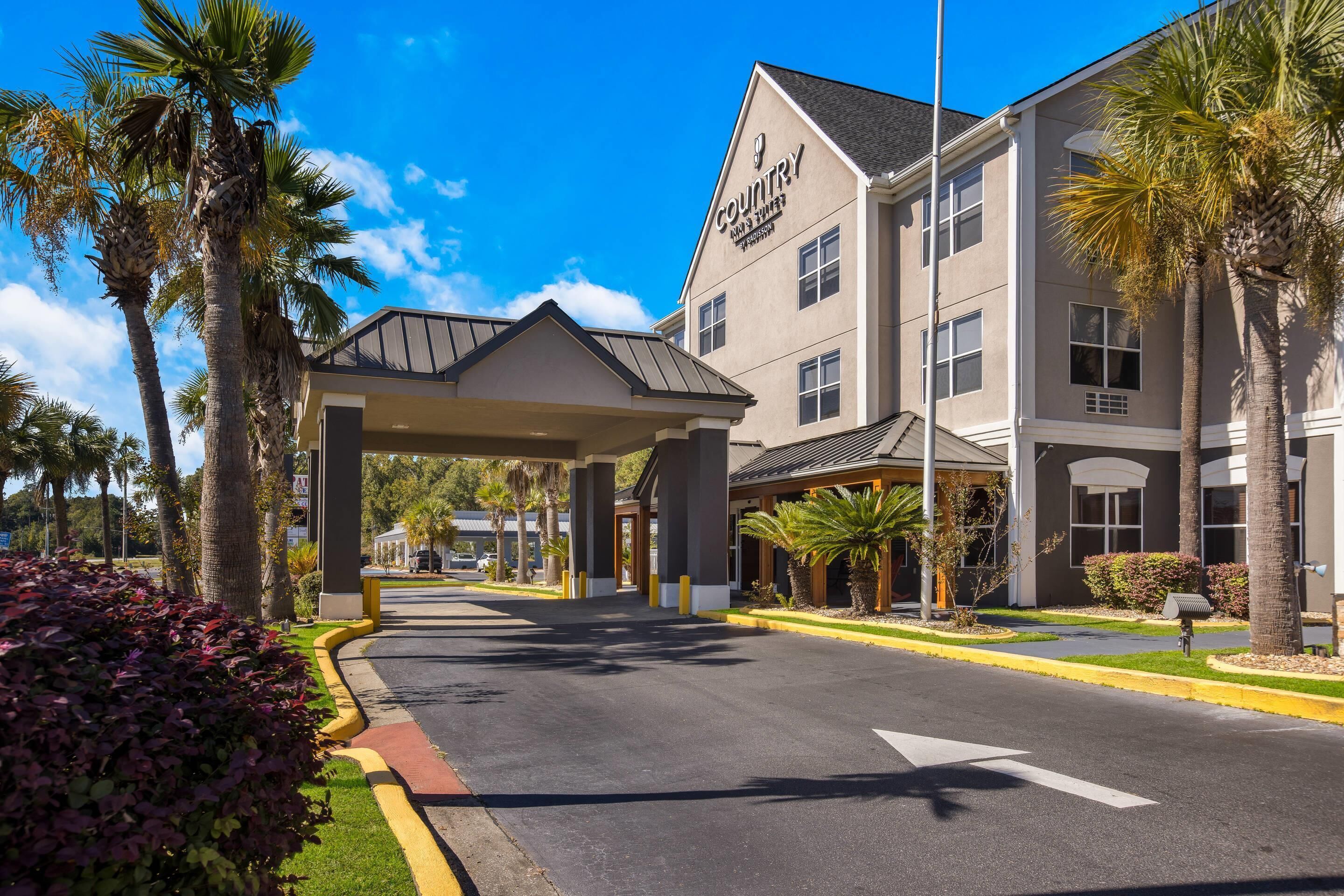 Entrada de hotel con un paseo techado, palmeras y cielo despejado.
