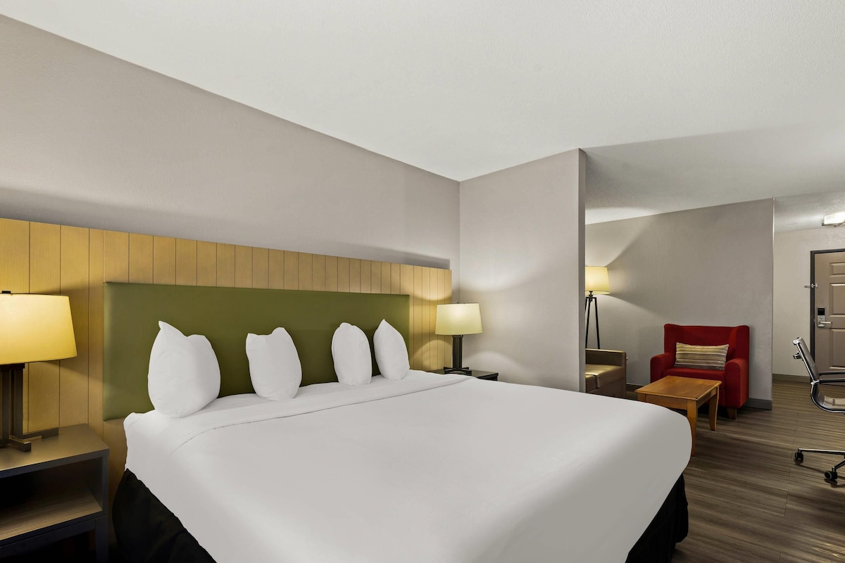 Habitación de hotel con una cama grande, dos mesitas de noche con lámparas, un sillón rojo, una mesa de centro de madera y una puerta con picaporte.