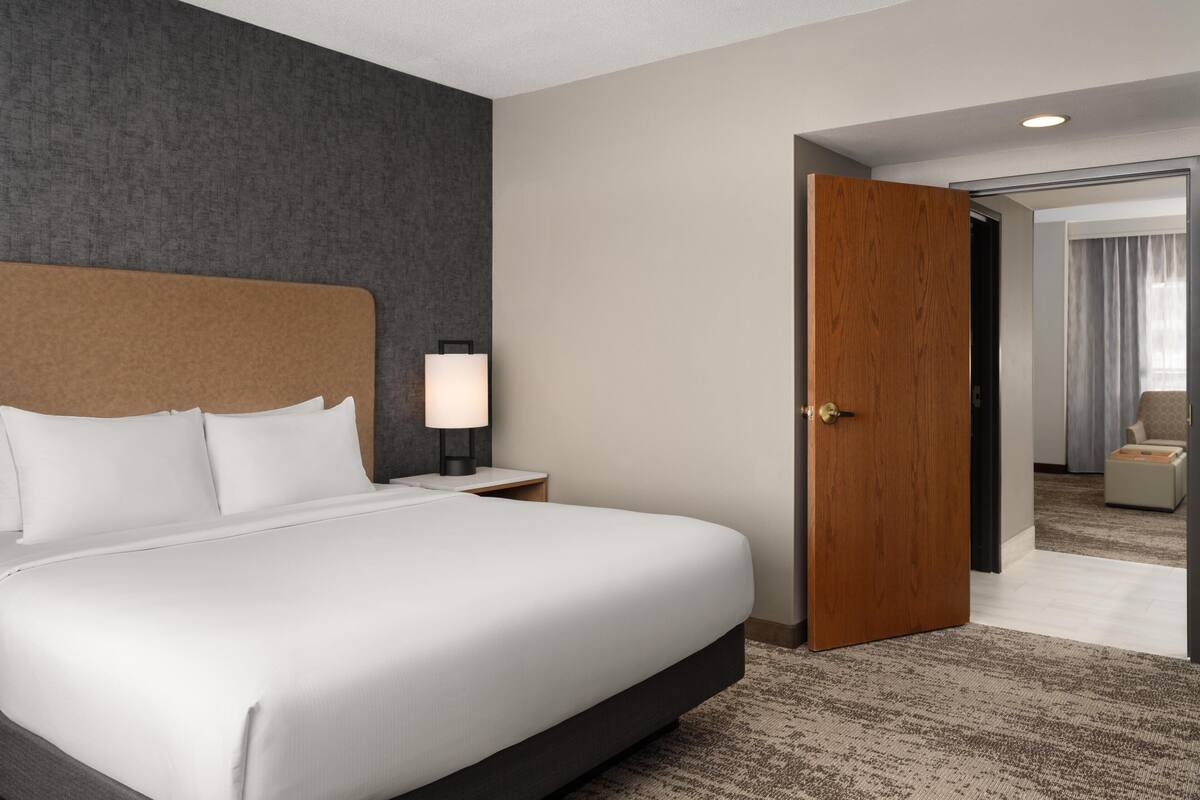 Ein Hotelzimmer mit einem großen Bett, einem Nachttisch mit Lampe, einer Holztür und Blick in ein weiteres Zimmer.