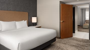 A hotel room with a large bed, a nightstand with a lamp, a wooden door, and a view into another room.