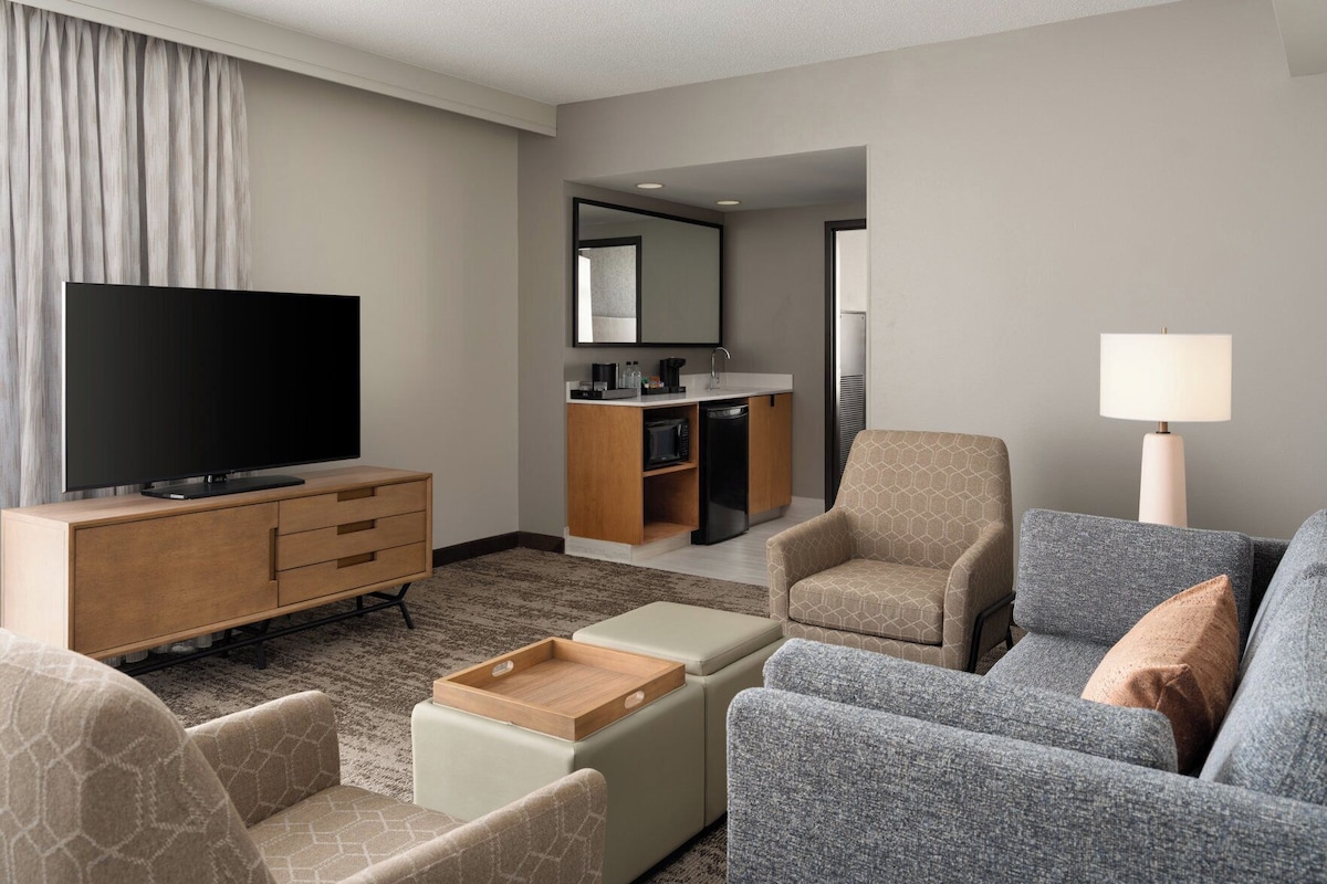 Une chambre d’hôtel équipée d’une télévision, d’un canapé, d’un fauteuil, d’un pouf et d’une kitchenette.