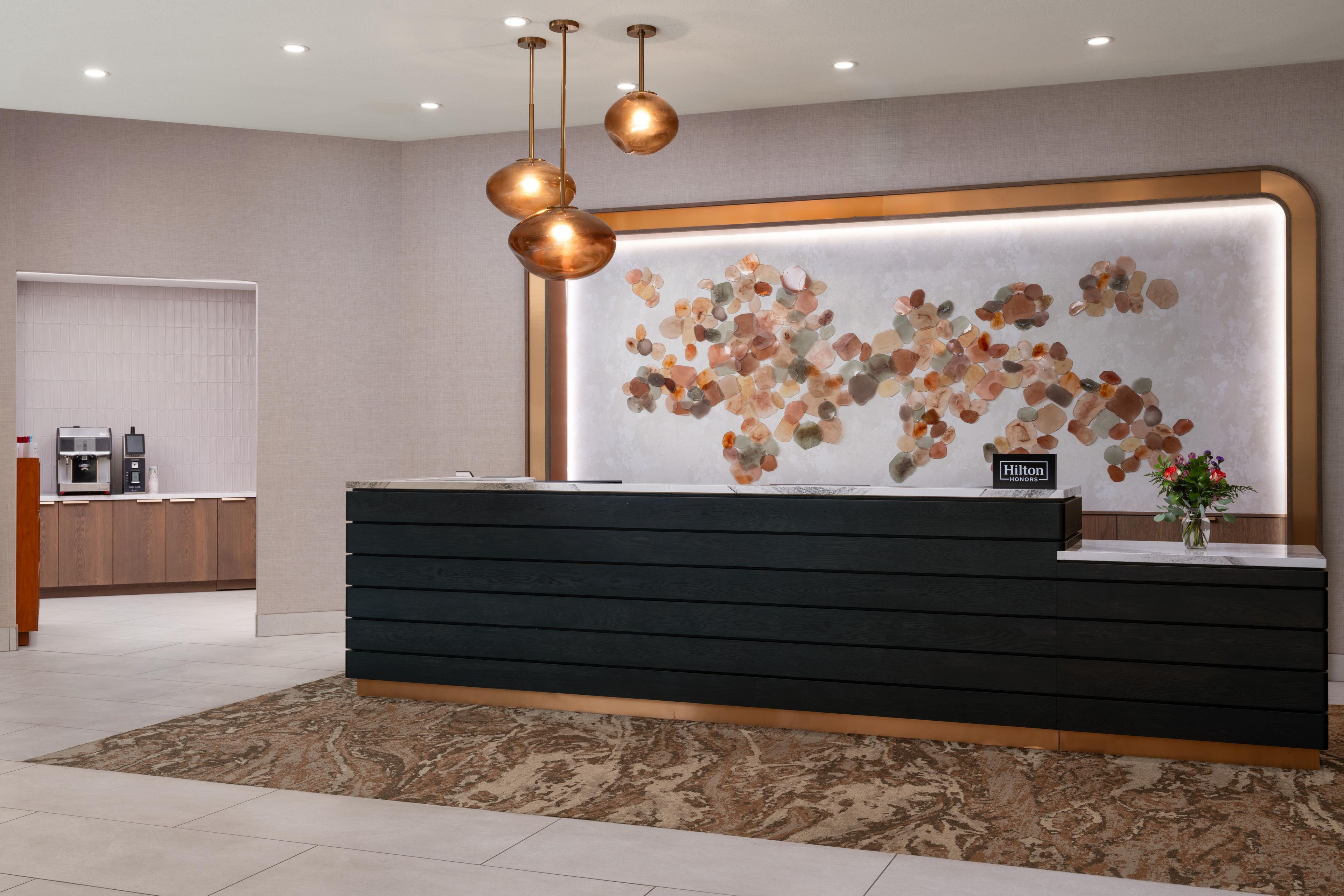 A hotel reception desk with a modern design, a wall-mounted artwork, and a coffee machine in the background.