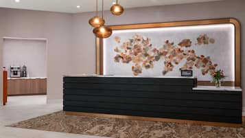 A hotel reception desk with a modern design, a wall-mounted artwork, and a coffee machine in the background.