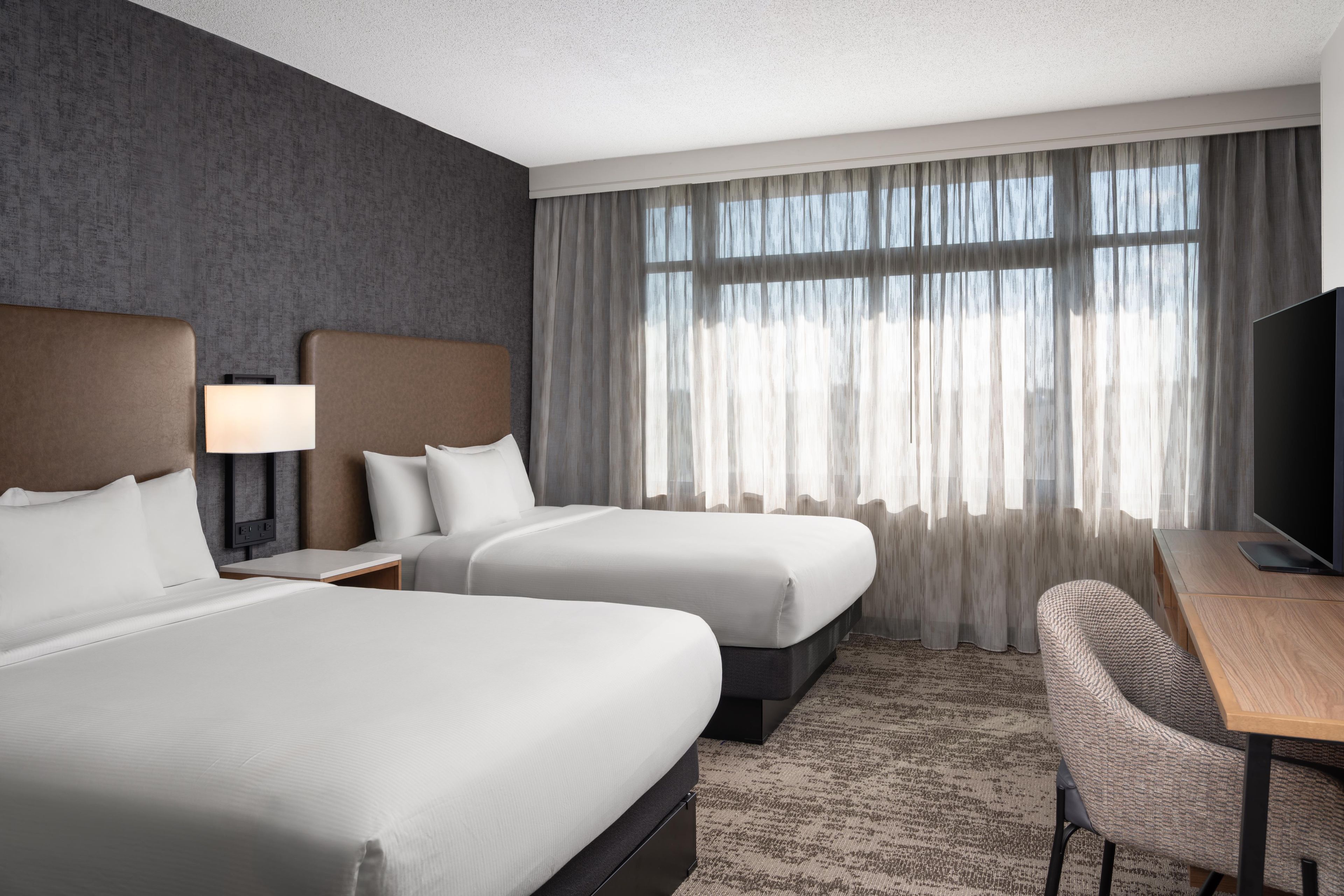 A hotel room with two beds, a desk, a chair, a television, and a large window with curtains.