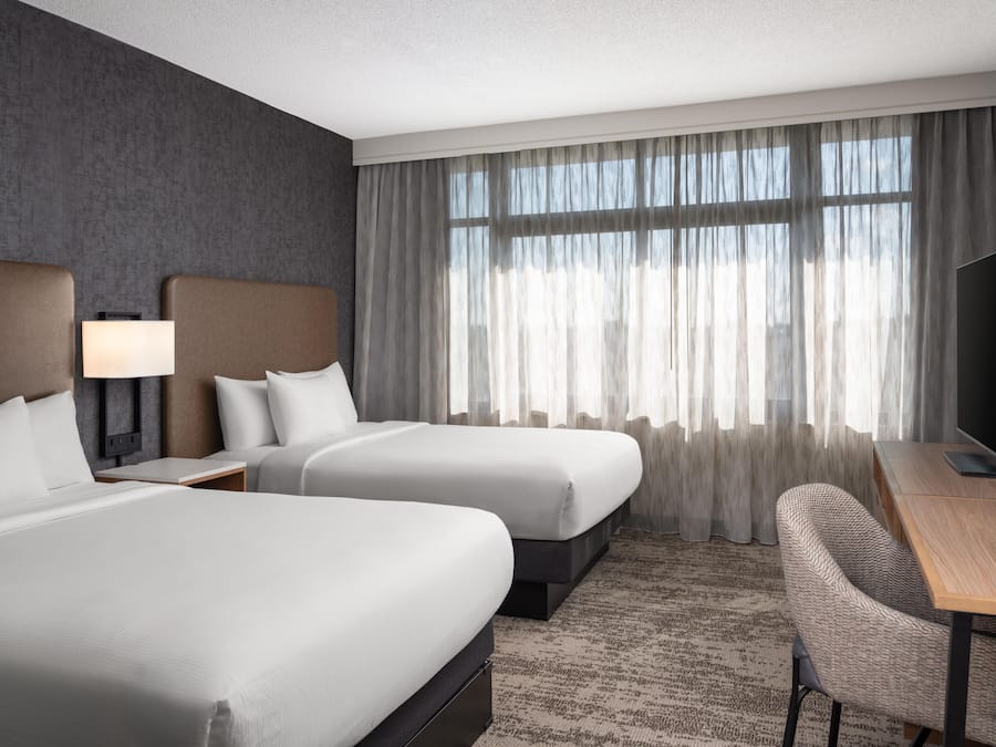 Ein Hotelzimmer mit zwei Betten, einem Schreibtisch, einem Sessel, einem Fernseher und einem großen Fenster mit Vorhängen.