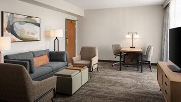 Habitación de hotel con sofá, sillón, reposapiés, escritorio, silla, televisor, lámpara y obras de arte.