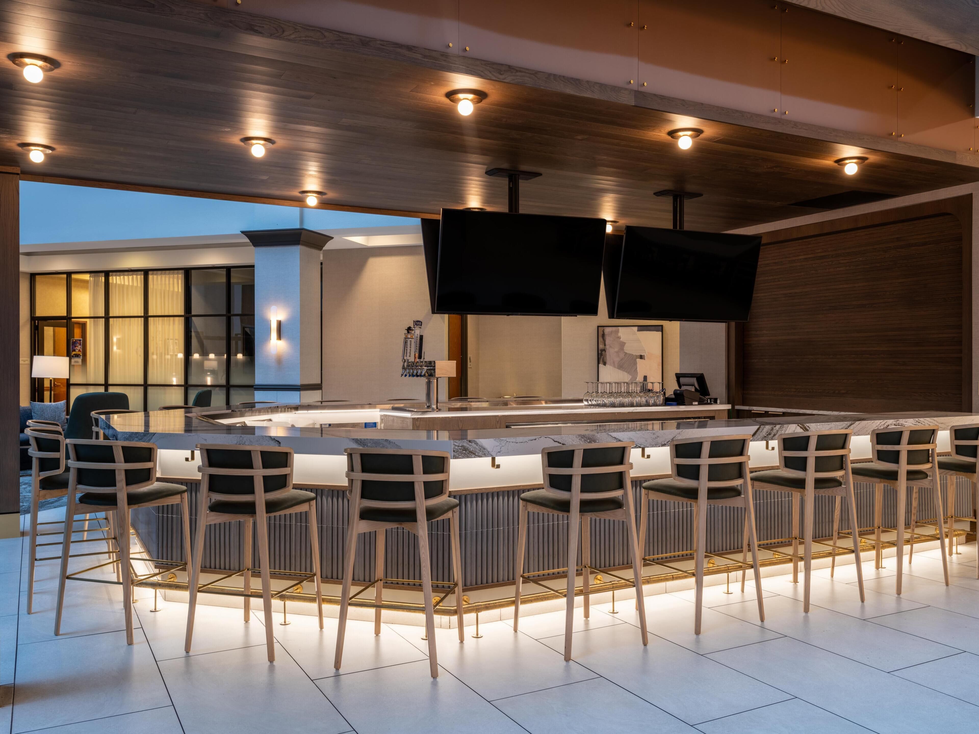 A modern bar area with a long counter, multiple bar stools, and a flat-screen TV mounted on the wall.
