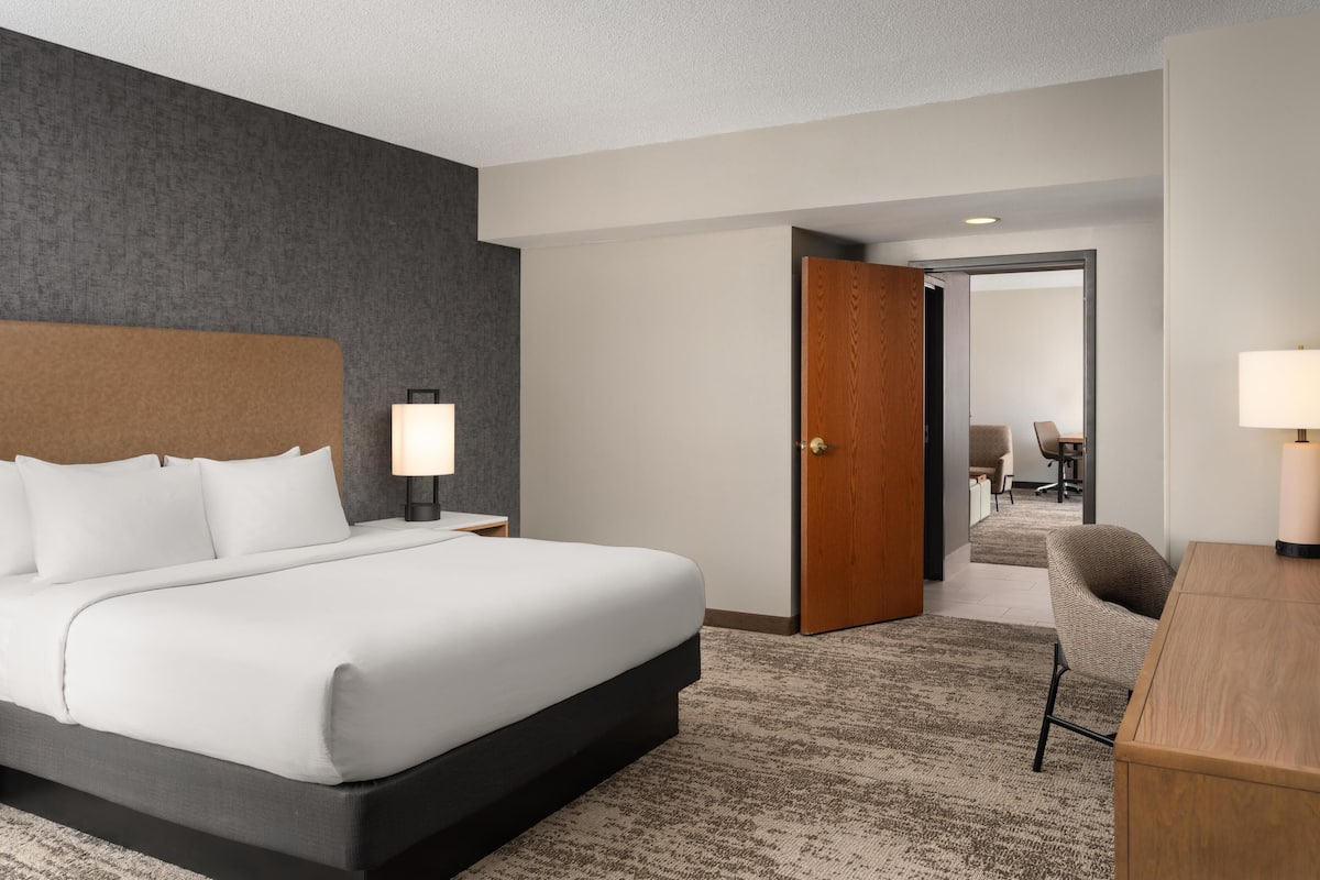 Ein Hotelzimmer mit einem großen Bett, einem Schreibtisch, einem Sessel, einem Nachttisch mit Lampe und einer Tür zu einem anderen Raum.
