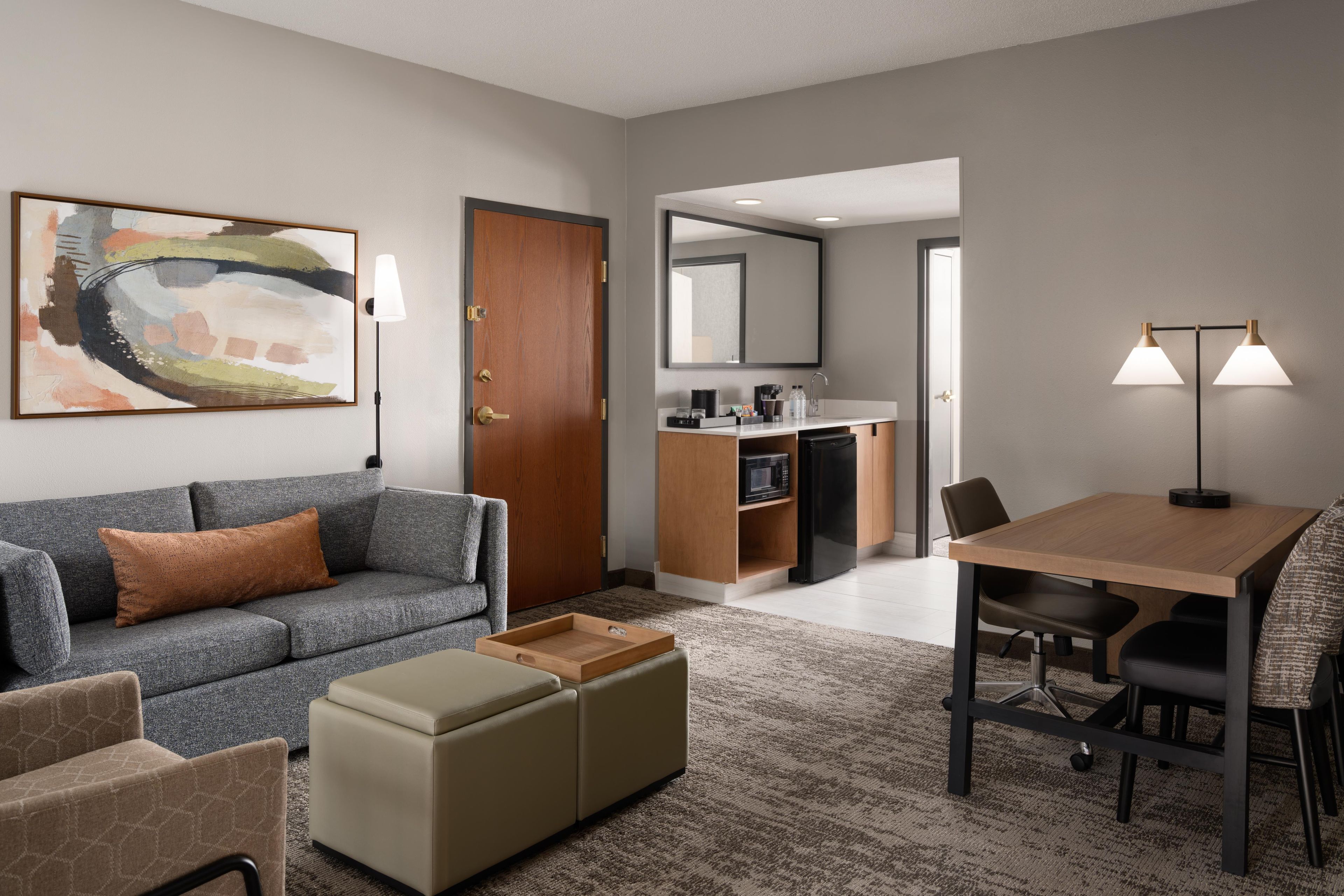 A modern hotel room with a gray sofa, a wooden dining table, a kitchenette, and a large abstract painting on the wall.