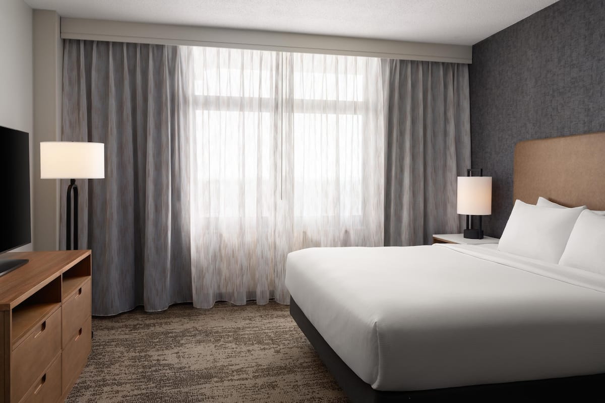 Ein Hotelzimmer mit einem großen Bett, zwei Nachttischlampen, einem Fernseher und einem Fenster mit Vorhängen.