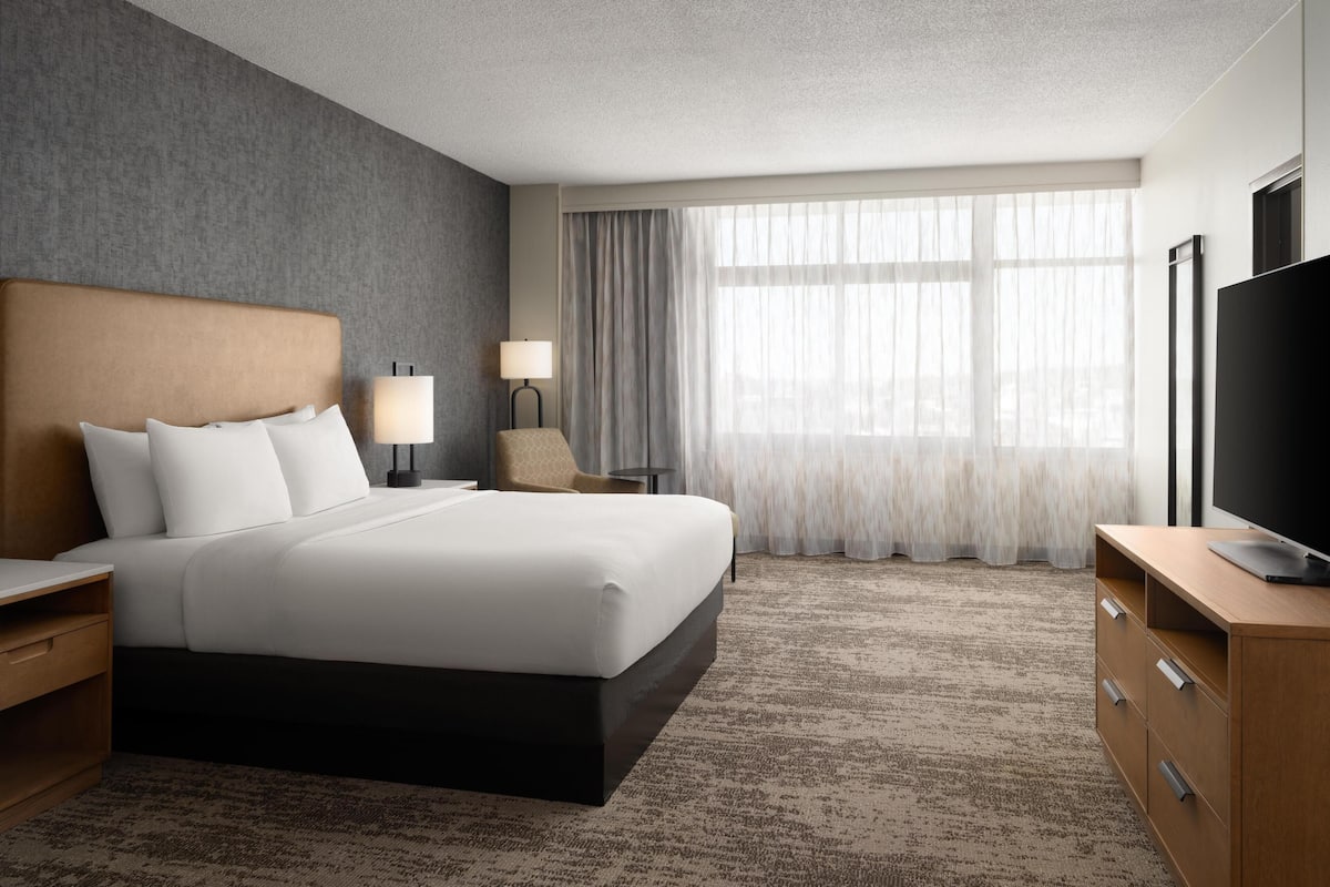 Ein Hotelzimmer mit einem großen Bett, einem Fernseher, einem Schreibtisch, einer Lampe und einem Fenster mit Vorhängen.