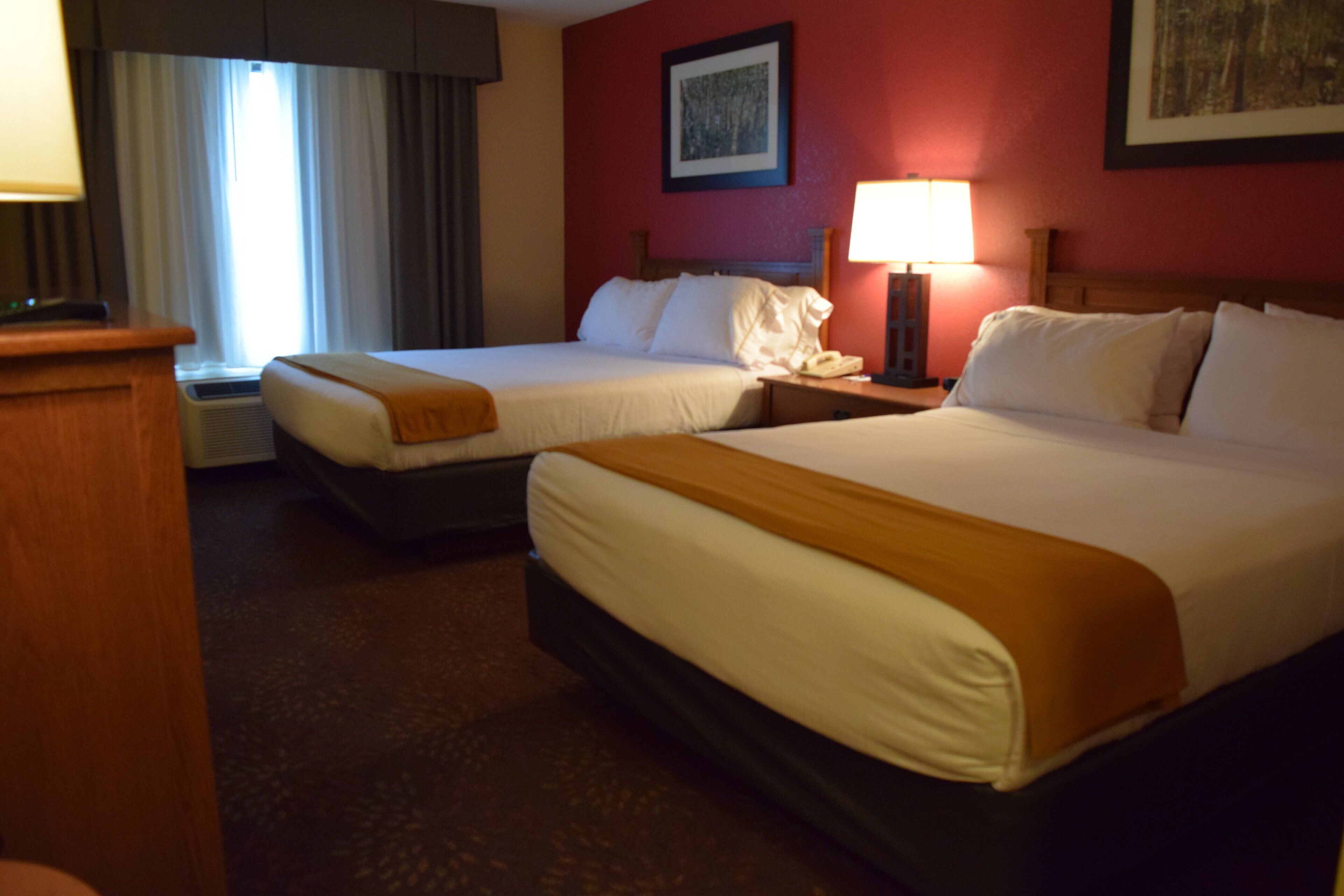 A hotel room with two beds, a nightstand, a lamp, and a window with curtains.