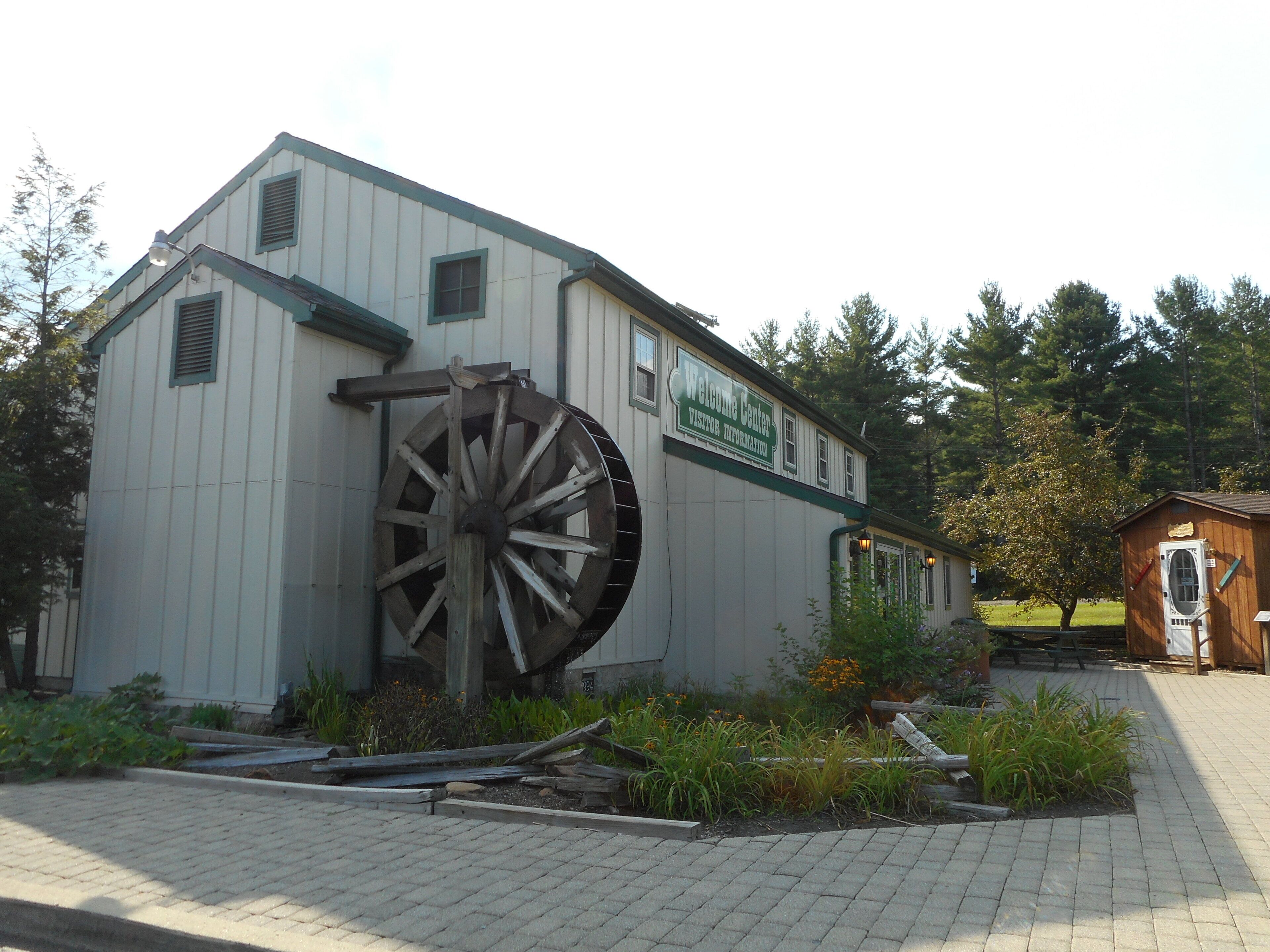A building with a large water wheel attached to its side, a signboard, and a small wooden structure nearby.